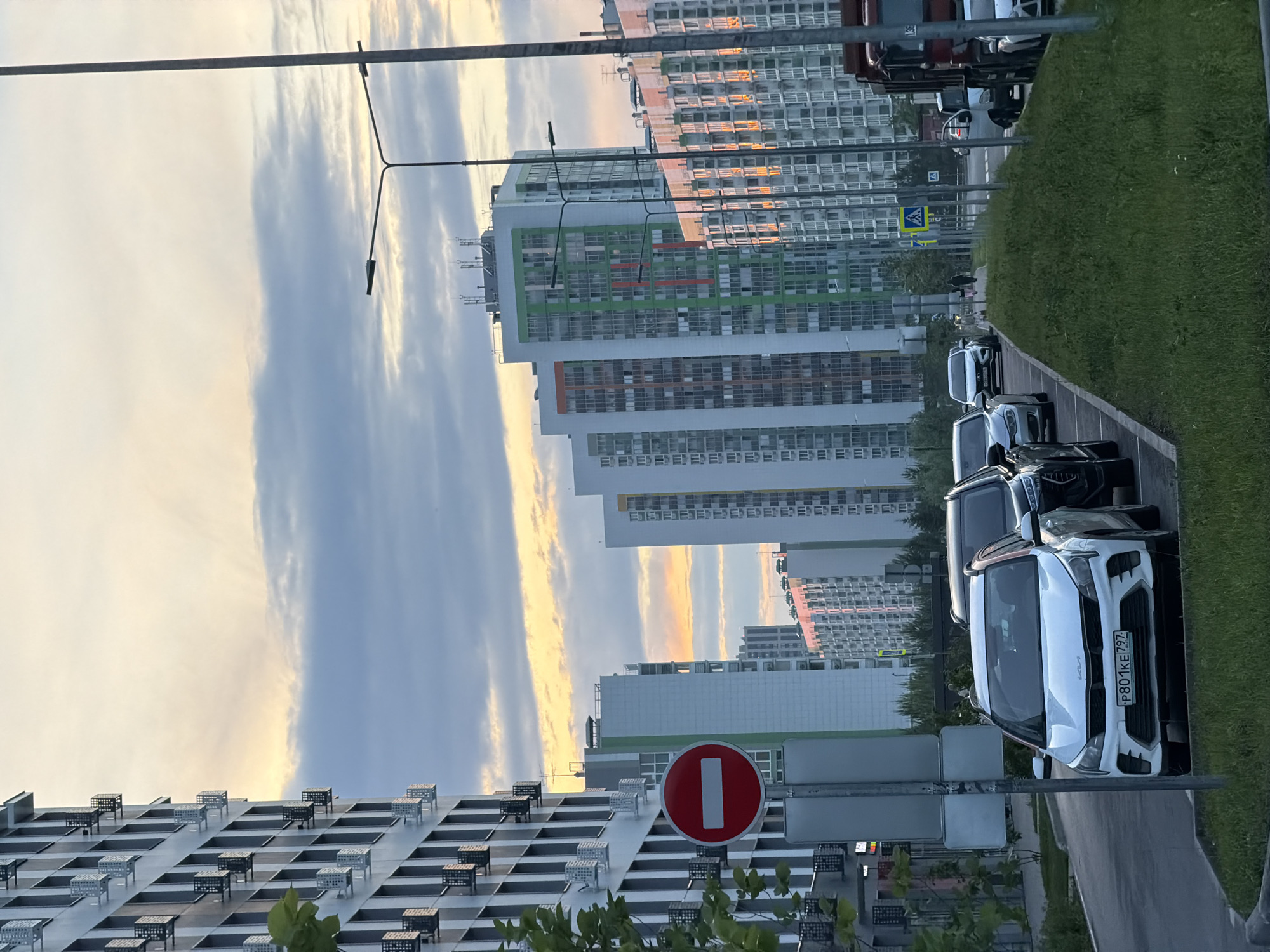 This image shows a street scene in a city with multiple high-rise apartment buildings under a cloudy sky. In the foreground, several cars are parked along the side of the road, including a white Kia with a license plate starting with "P801KE". A red and white "no entry" sign is visible on a pole to the left of the cars. The apartment buildings are modern with numerous windows and some feature colorful accents. Streetlights line the road. The lighting suggests it is either dawn or dusk, with soft, warm light visible on some of the building facades. A red truck is parked further down the street.