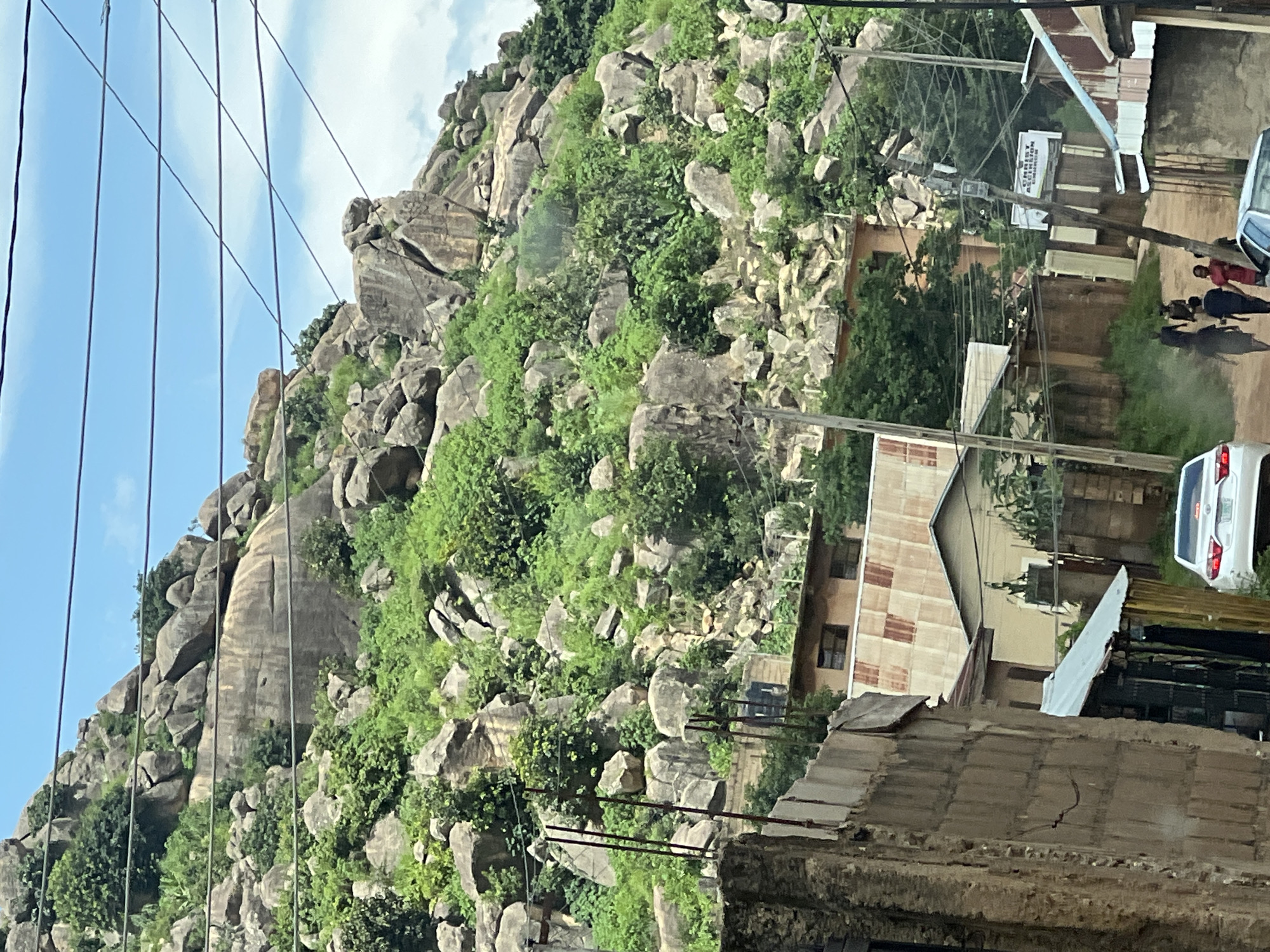 The image depicts a view of a rocky hillside covered in lush green vegetation, with a few buildings nestled at its base. In the foreground, there's a dirt road leading into the scene, with a white car parked on the right. Several individuals are walking on the road, some appearing to be young men and children. The background is dominated by the expansive, boulder-strewn hillside, with power lines crisscrossing the sky. Several buildings, some with corrugated iron roofs, are visible amongst the trees and rocks. One building has a sign that reads "CHRIST ASCENSION CHURCH". The overall impression is a blend of natural landscape and human habitation in a somewhat rugged environment.