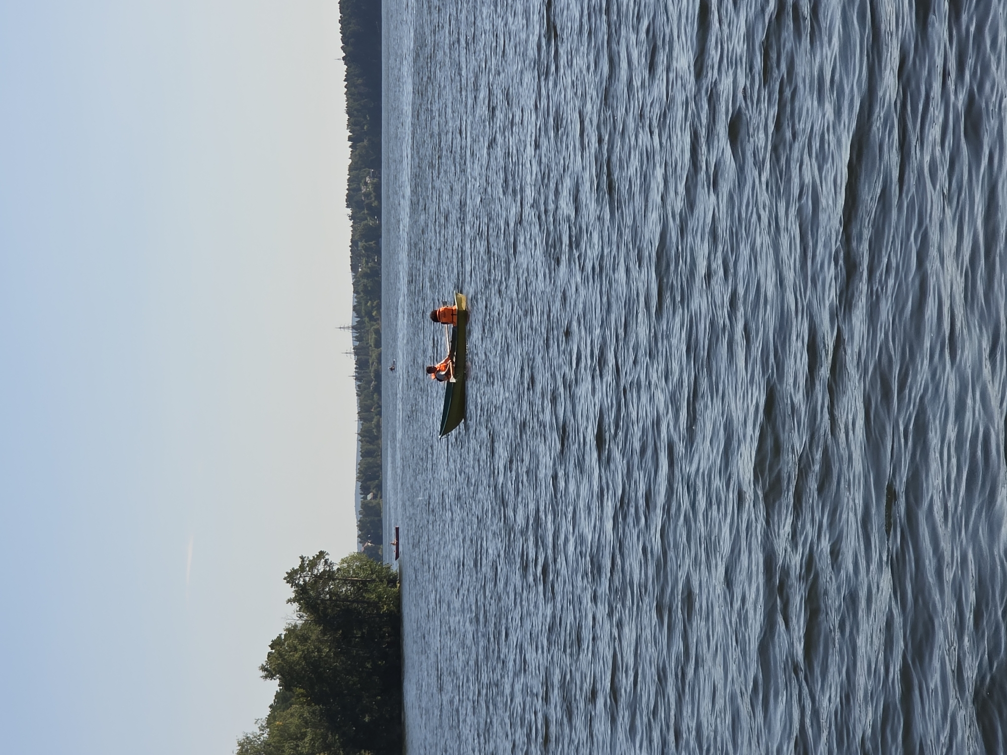 In Chebarkul, Russia, on a body of water, a small green rowboat is depicted with two individuals. The individuals are positioned towards the front of the boat, both wearing bright orange life vests. One individual, appearing to be an adult, is seated and facing forward, while the other, possibly a child, is seated behind them, also facing forward. The boat is on a wide expanse of blue water, characterized by moderate ripples and waves. In the distance, a treed shoreline with scattered buildings and industrial structures, including what appear to be power line towers, is visible. The sky is clear and pale blue.