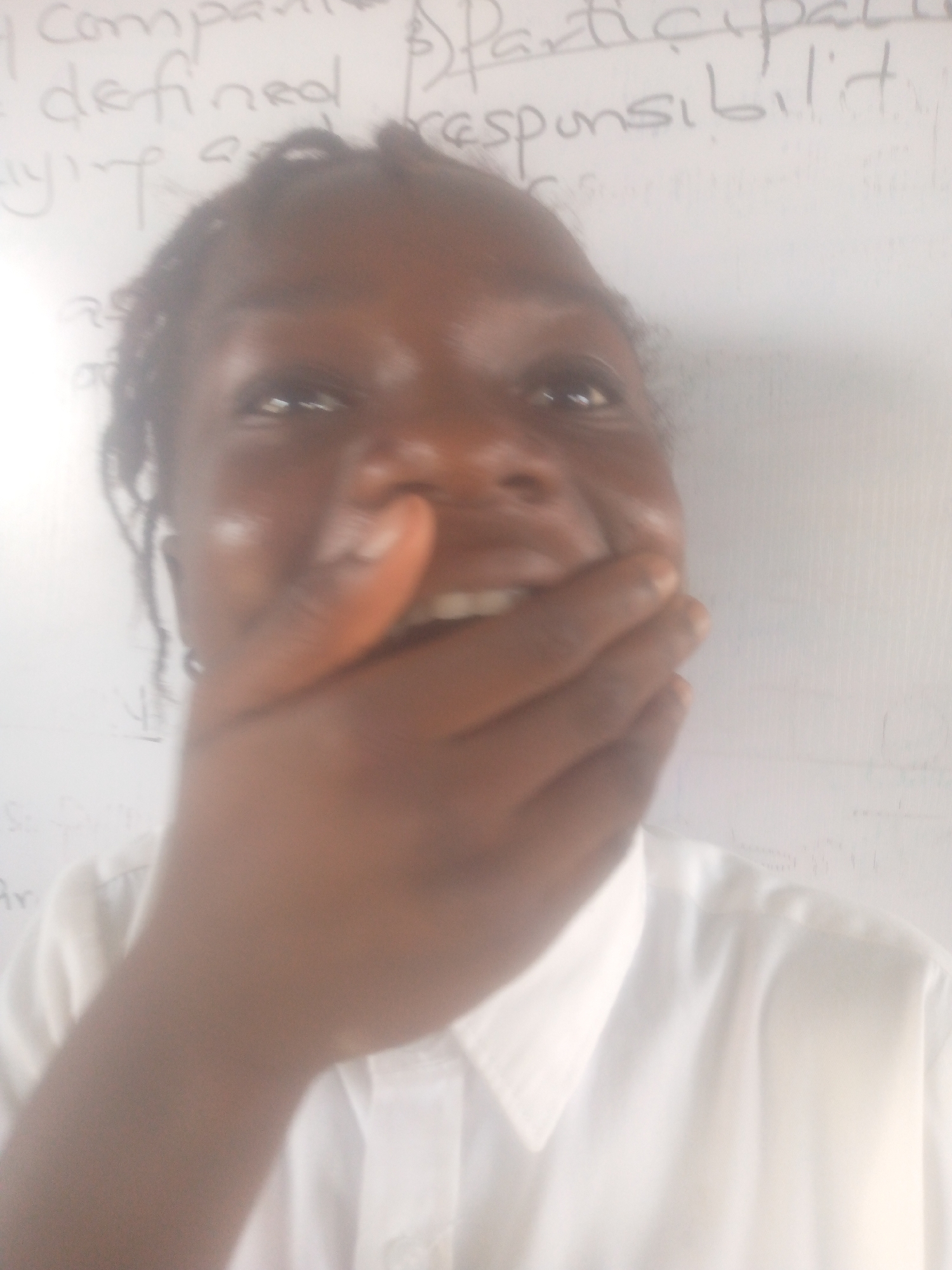 A person with dark skin is covering their mouth with their hand, possibly laughing or surprised. They are wearing a white collared shirt. In the background is a white board with writing, including the word "responsibility". The location and city are unknown.