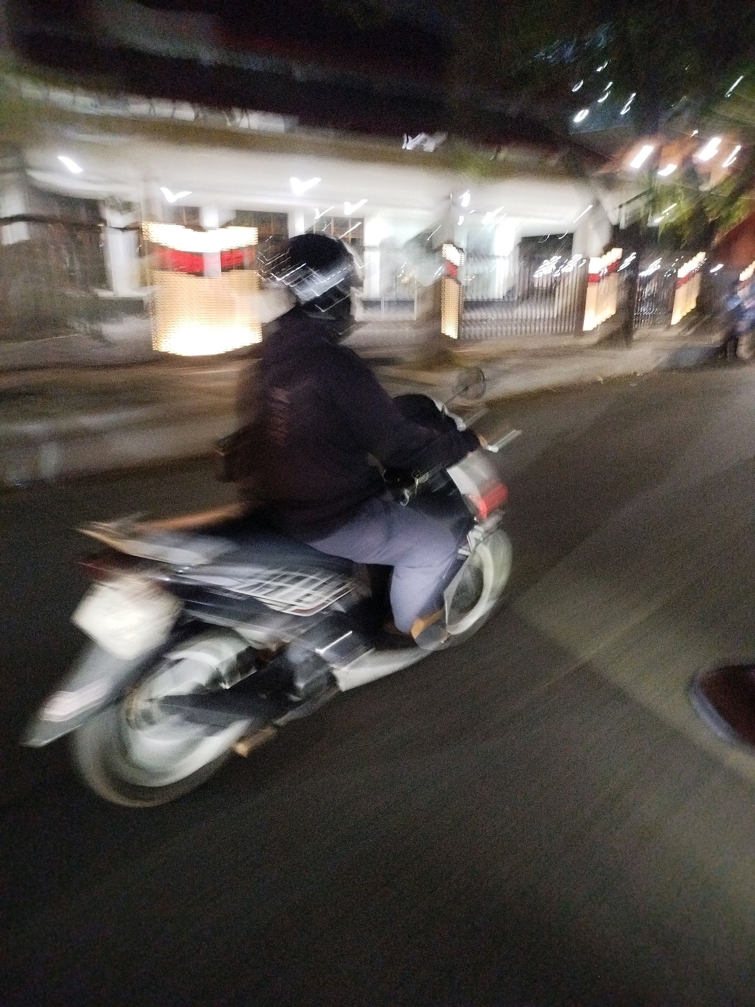 This is a motion blurred, night time photograph taken from a moving vehicle, likely a car, on a city street in Surabaya, Indonesia. The primary subject is a person riding a scooter, viewed from the side and slightly from behind. The rider is wearing a dark hooded jacket and a dark helmet with a visor that obscures their face. They are wearing grey pants and sandals. The scooter is silver and black with visible wheels and a rear fender. The background shows the blurred outlines of buildings with illuminated windows and decorative lighting. The street itself is dark asphalt, with some lighter areas indicating reflections or streetlights. The overall impression is one of movement and low light conditions, characteristic of a night scene in a bustling urban environment.