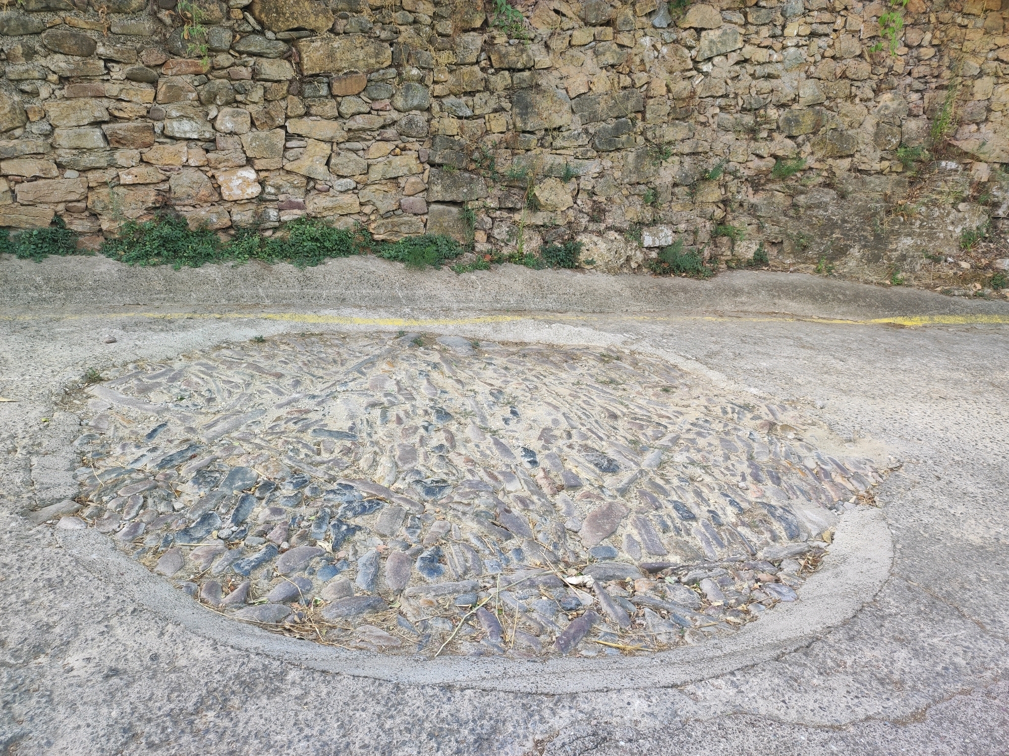 The image shows a cobblestone circle in the city of Sueras, Spain. The circle is made of dark, irregularly shaped stones and is surrounded by concrete. A yellow line runs across the concrete surface behind the cobblestone circle. Behind the concrete, a stone wall with plants growing at its base stretches across the frame. The wall is constructed from rough, uneven stones of various sizes and colors, with some moss and vegetation clinging to it. The ground in front of the wall is a mixture of concrete and the cobblestone circle. The overall scene depicts an outdoor area, likely a street or plaza, with a rustic and aged aesthetic.