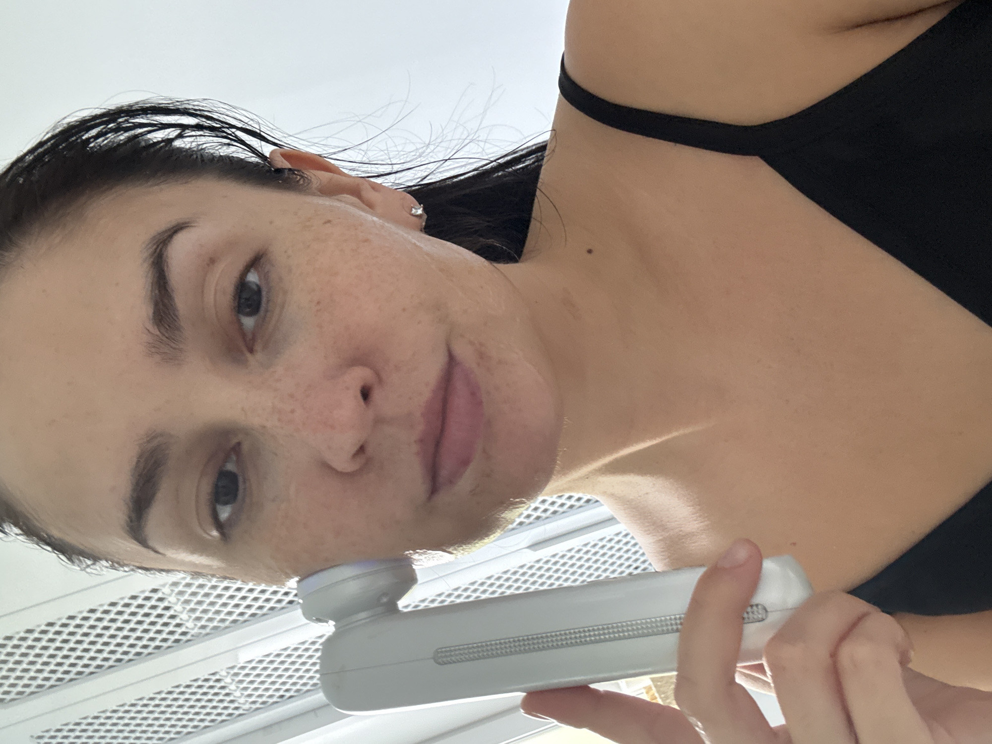 A woman is holding a white facial cleansing device to her cheek and the side of her face. She has dark hair pulled back, brown eyes, and freckles. She is wearing a black tank top and a small stud earring in her right ear. The background appears to be an indoor setting with a white, slatted structure on the left side of the image and a plain white wall on the right. There is no information in the image to suggest a location in Nizhneyarkeyevo, Russia.