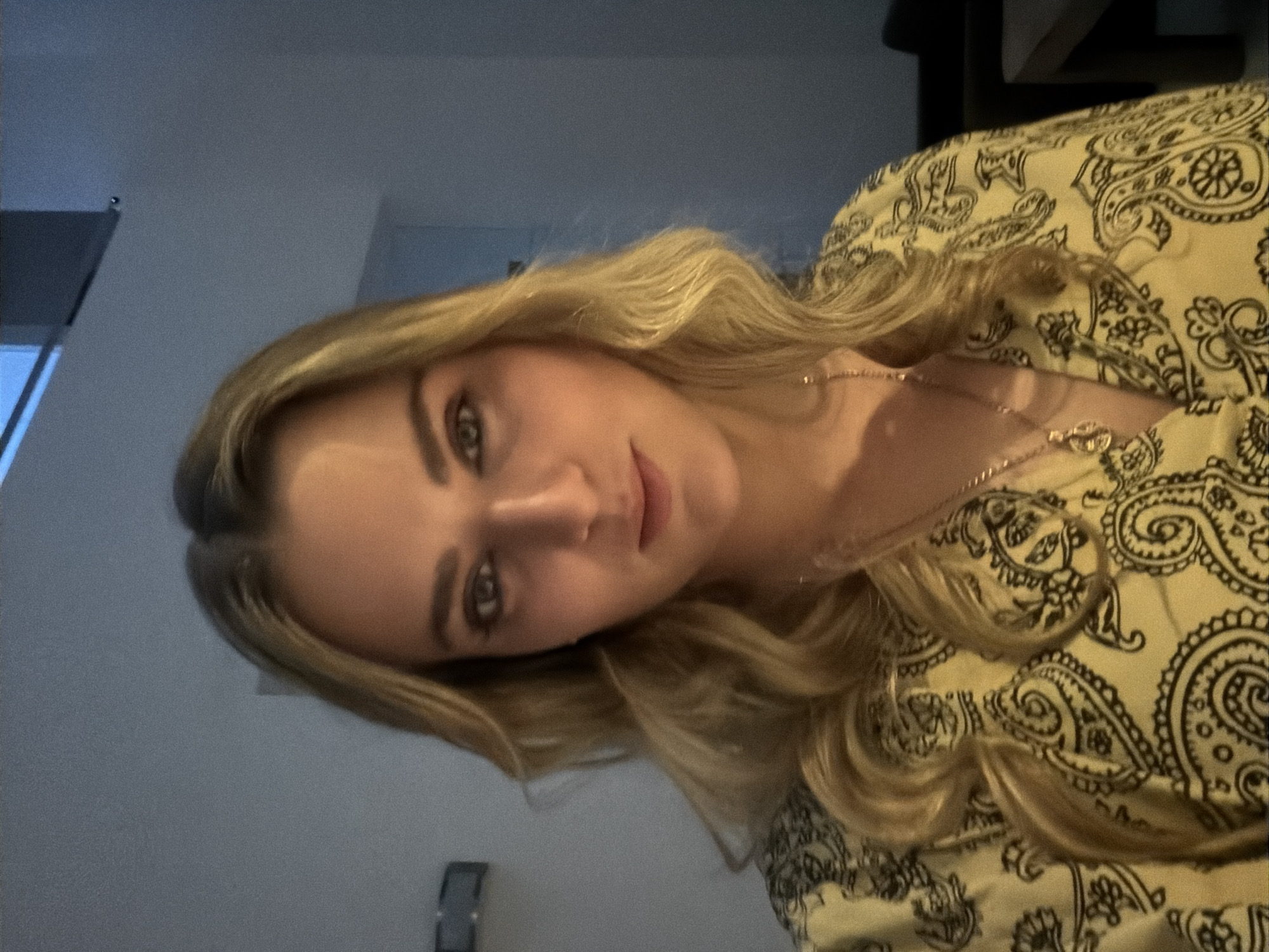 A medium shot depicts a blonde woman wearing a light-yellow, paisley-patterned blouse and a delicate gold necklace. The background includes a light gray wall and a glimpse of a window to the upper left. There is no visual information indicating the image was taken in San Bernardino Tlaxcalancingo, Mexico.