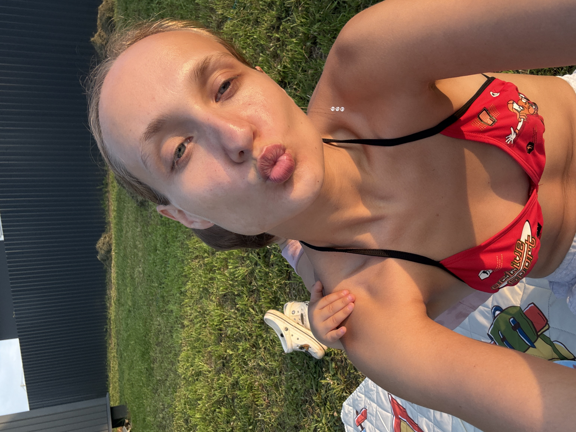 A young woman with blonde hair is lying on the grass, taking a selfie. She is wearing a red bikini top with cartoon characters and black straps. A small necklace with three white beads is visible on her neck. Her lips are pursed in a kiss. Beside her is a white Croc shoe and a blanket with a cartoon dinosaur print. In the background, a dark, ribbed structure and green grass are visible. The lighting suggests it is late afternoon or early evening, with the sun casting a warm glow. The scene appears to be outdoors, in a grassy area, possibly a backyard or park. No specific event is indicated, other than the woman relaxing and taking a photo. The text "ACTIVE SPORT" is visible on her bikini top.