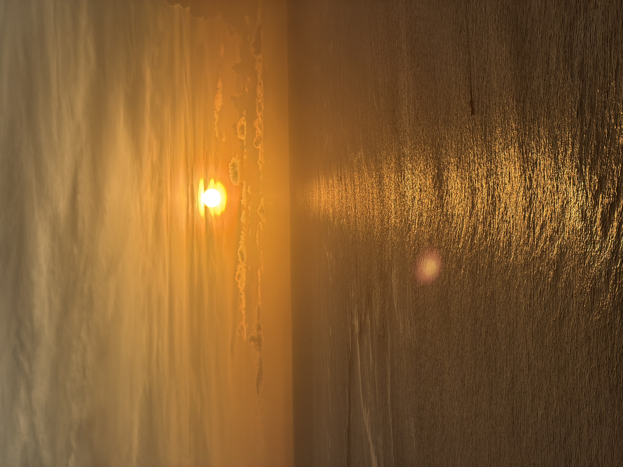 The image shows a seascape at sunset, likely in Kuta Selatan, Indonesia. The sun is setting over the ocean, with its bright glare reflected on the water's surface. The sky is filled with clouds, and the lighting is warm and golden. The foreground features the ocean water's surface, which appears textured due to the play of light.