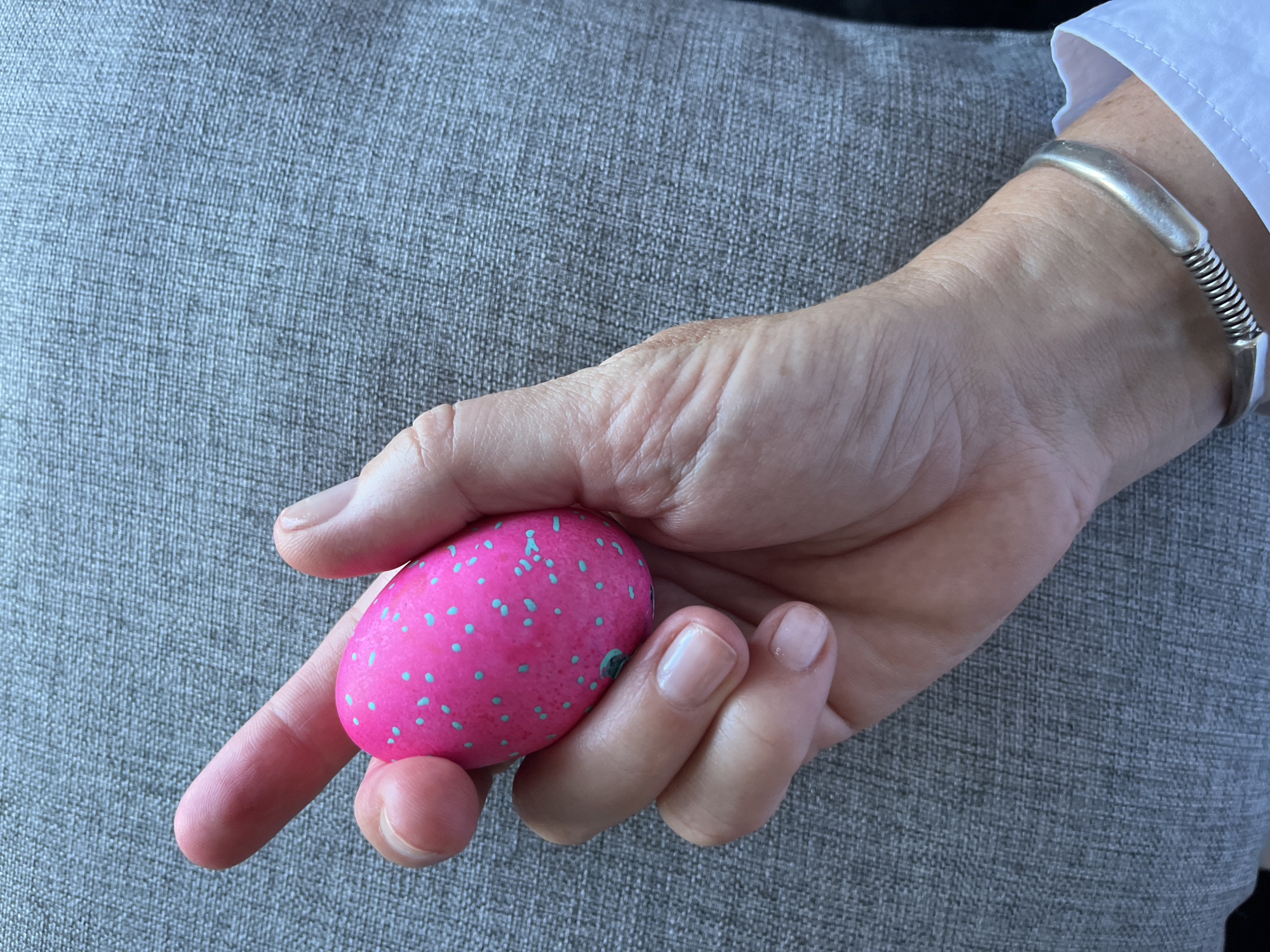 A hand is holding a pink Easter egg with light blue spots. The hand is wearing a silver bracelet. The background is a gray fabric. There is no indication of the location being Denpasar, Indonesia.