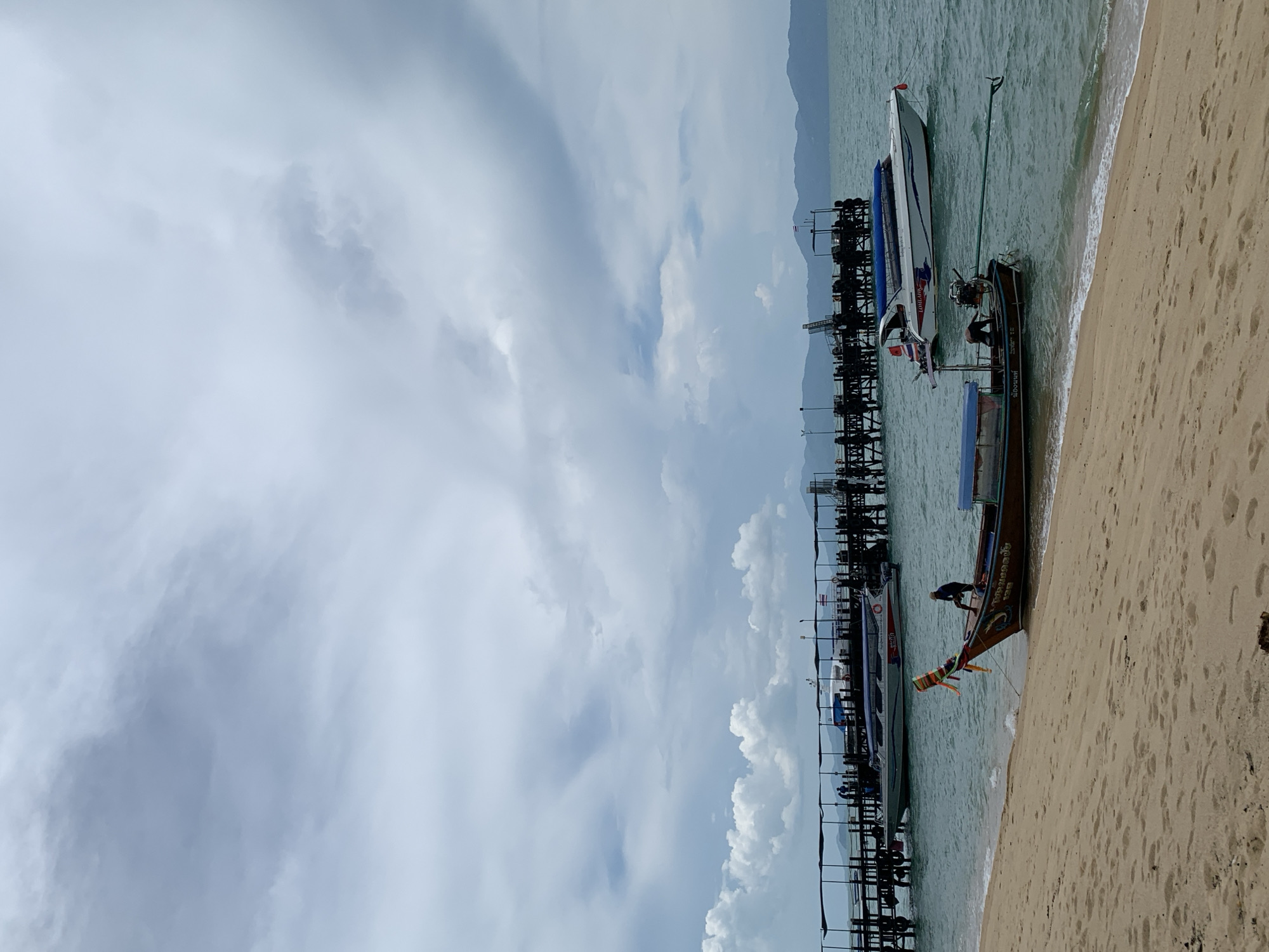 The image shows a beach scene in Baan Tai, Thailand, with boats docked near a pier. The sky is cloudy. One boat, a traditional longtail boat, is partially beached on the sandy shore. A person is visible on the longtail boat, performing some activity. A white speedboat is docked nearby. The pier extends into the water and has supports visible in the water.