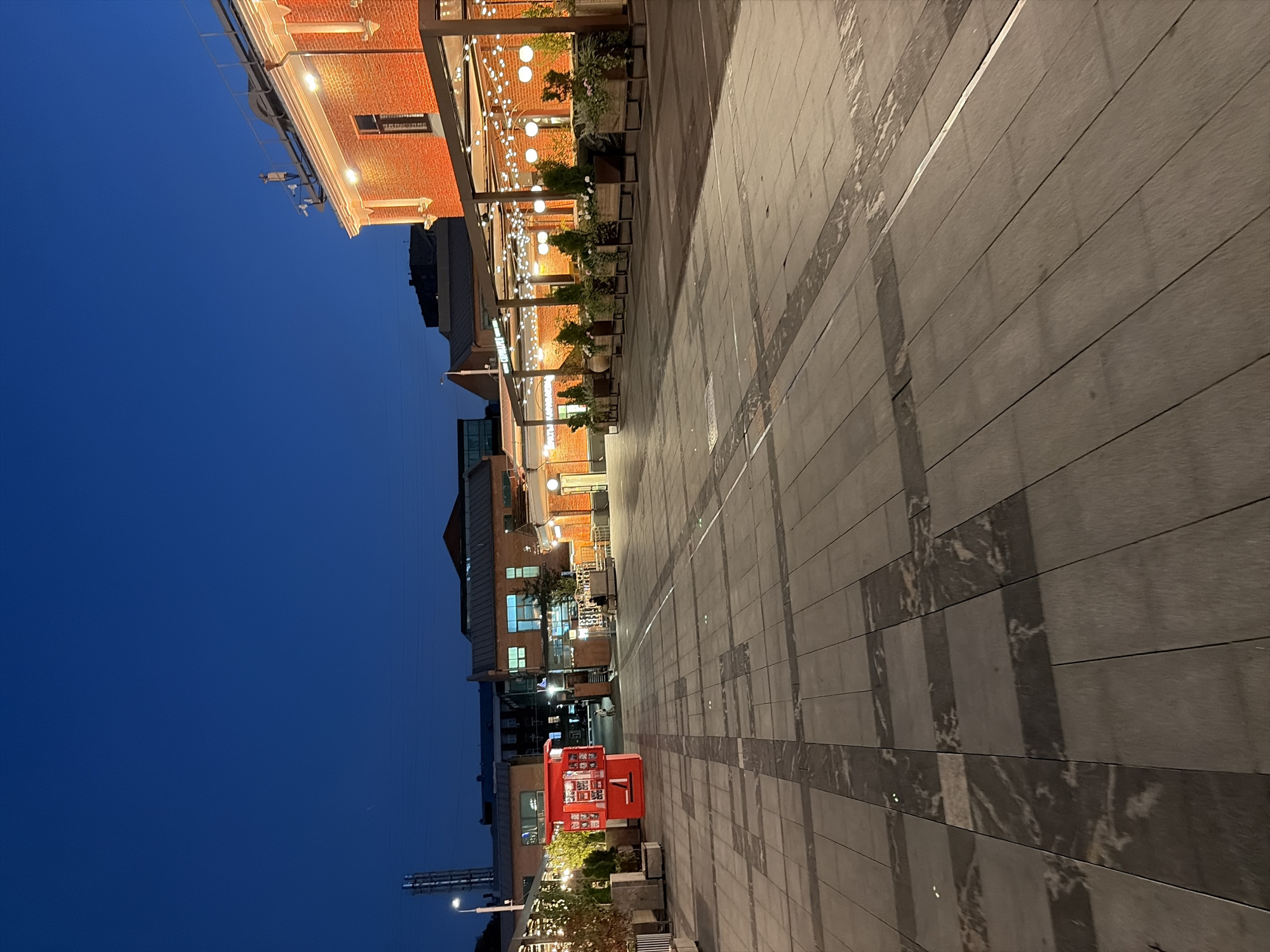 This is an evening photograph of a wide, paved outdoor area in Moscow, Russia, likely a pedestrian street or plaza. The sky is a deep, dark blue, indicating twilight or early night. The paving stones are a mix of gray tones with darker, marble-like veins, laid out in a grid pattern with some areas appearing lighter due to reflection or lighting.
On the left side of the scene, a bright red kiosk or small building stands out. Further back, a multi-story brick building with windows is visible. To the right, a long, red-brick building with decorative white trim is illuminated by warm light. Along the front of this building, there is a covered outdoor seating area for a cafe or restaurant, adorned with strings of white fairy lights and larger spherical lights. Planters with small trees and greenery are placed in front of the seating area.
A few scattered lights are visible on poles, and the general ambiance suggests a public space that is open for business or leisure, despite the late hour. There are a few people visible in the distance, including one person walking near the red kiosk and another sitting on a bench near the cafe area. The overall scene is clean, modern, and well-lit, creating a welcoming atmosphere.