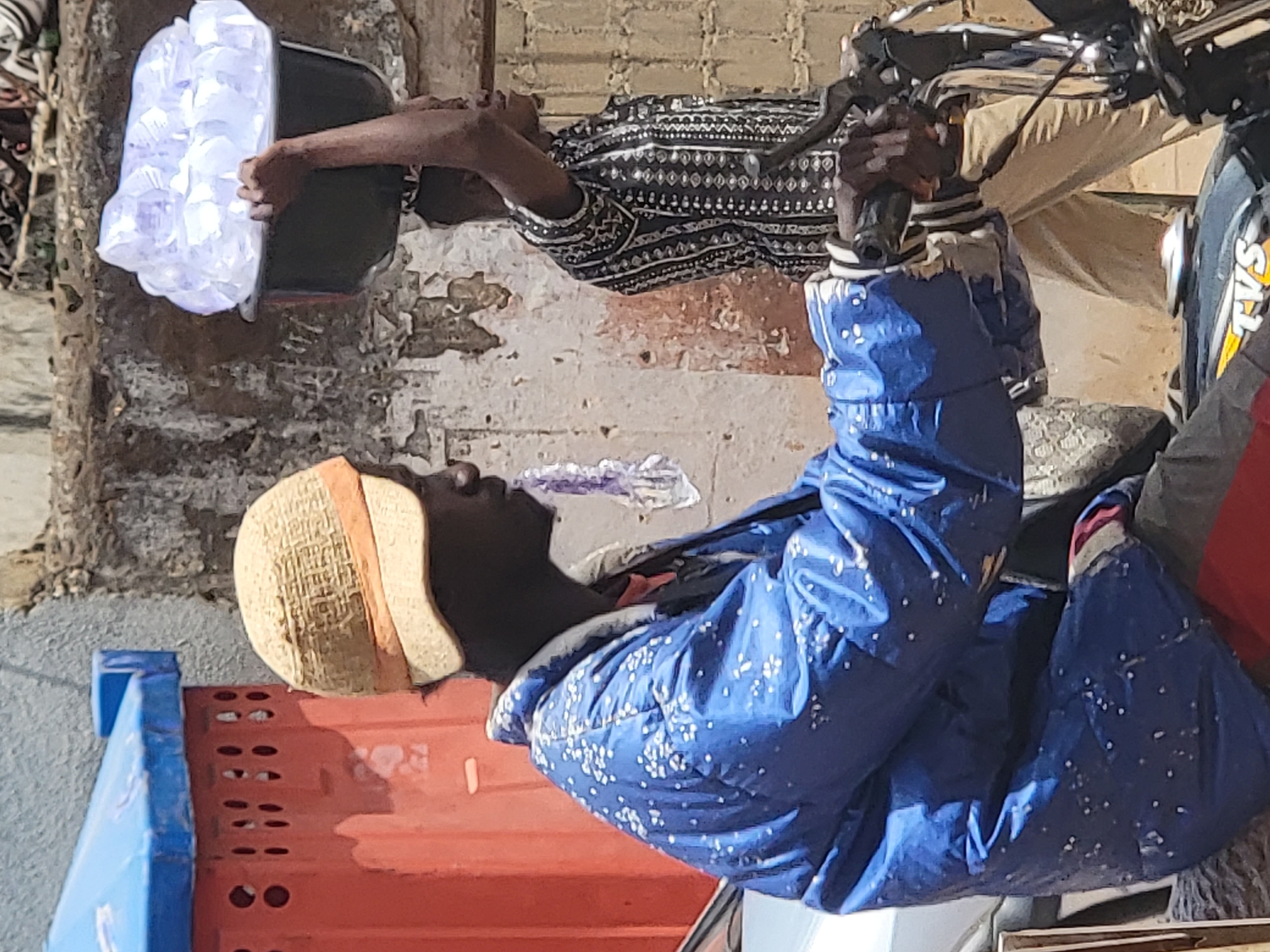 A man in a blue jacket and straw hat sits on a motorcycle in Maiburiji, Nigeria. Another man stands nearby, holding a bucket filled with translucent plastic bags. A blue and red barrier is visible in the foreground.