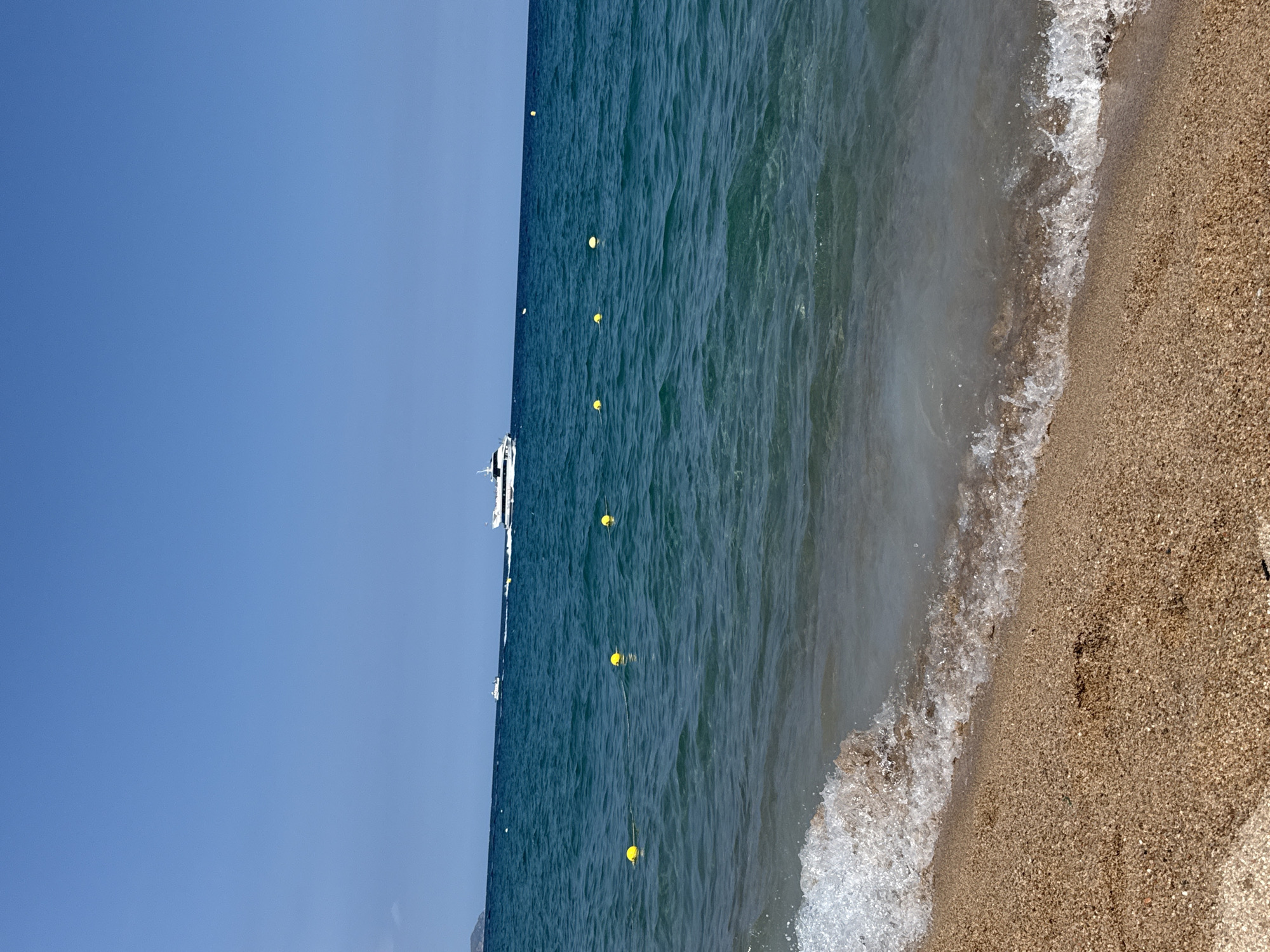The image depicts a sunny day at a beach in Blanes, Spain. The foreground features a sandy shoreline with a gentle wave breaking. The sea is a clear turquoise color, with several yellow buoys floating on the surface, indicating a designated swimming area. In the distance, a white boat with multiple decks is sailing across the water, leaving a faint wake. The sky is a bright, cloudless blue. The overall scene conveys a sense of tranquility and summer vacation.
