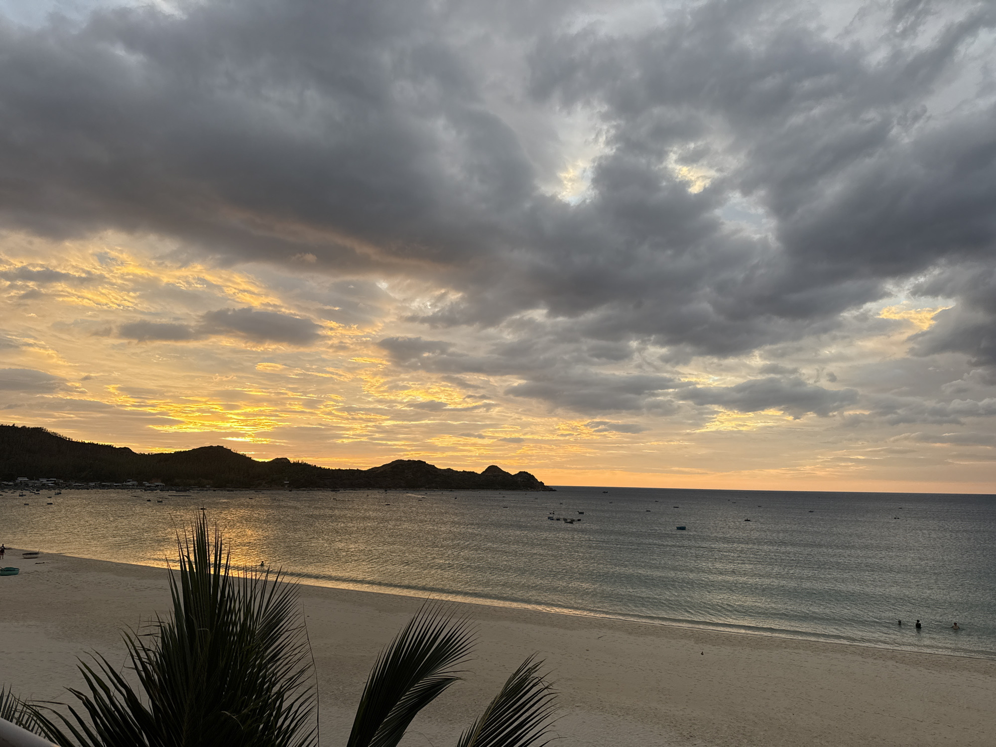 The image depicts a sunset over Bai Xep beach in Song Cau, Vietnam. The sky is filled with dramatic, heavy grey clouds interspersed with bright, warm yellow and orange hues from the setting sun. The sea is calm and reflects the colorful sky. In the distance, a coastline with hills and a small village is visible. Several small boats are scattered across the water. The foreground features a sandy beach with a few people visible, and the fronds of a palm tree partially obscure the view.