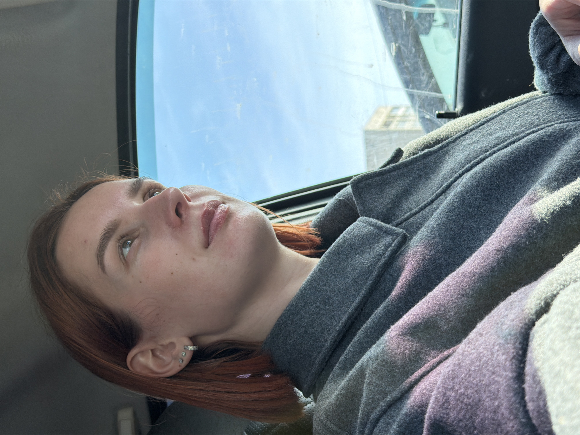 The image depicts a woman with auburn hair wearing a grey wool coat in what appears to be a car, looking upward towards the sky visible through the car window. The location is Chaikovsky, Russia, as evidenced by the building visible through the window. She has a fair complexion and multiple earrings in her left ear. The sky is blue and mostly clear with some clouds. The interior of the car is visible.