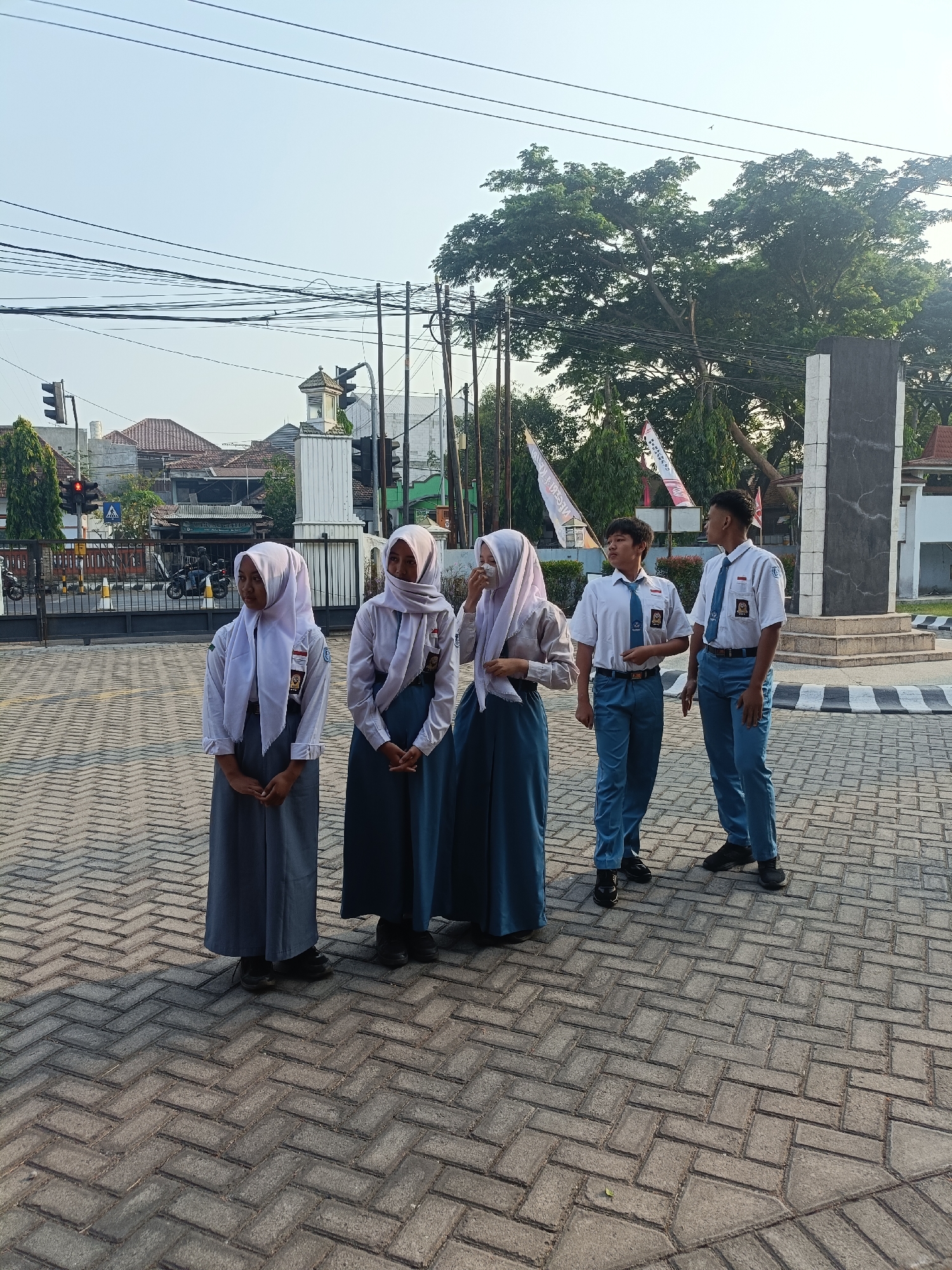 In Kediri City, Indonesia, five students in school uniforms stand on a paved area with a herringbone pattern. The three female students on the left wear white long-sleeved shirts, grey skirts, and light purple headscarves. The two male students on the right are dressed in white short-sleeved shirts, blue ties, and blue pants. The background reveals a street scene with a traffic light, buildings with tiled roofs, a fence, and several trees, including a large one with lush green foliage. A dark, rectangular monument with a grey facade stands to the right of the students. Power lines crisscross the sky, and two flags with Indonesian text are visible behind the students. The scene appears to be an outdoor school gathering or event.