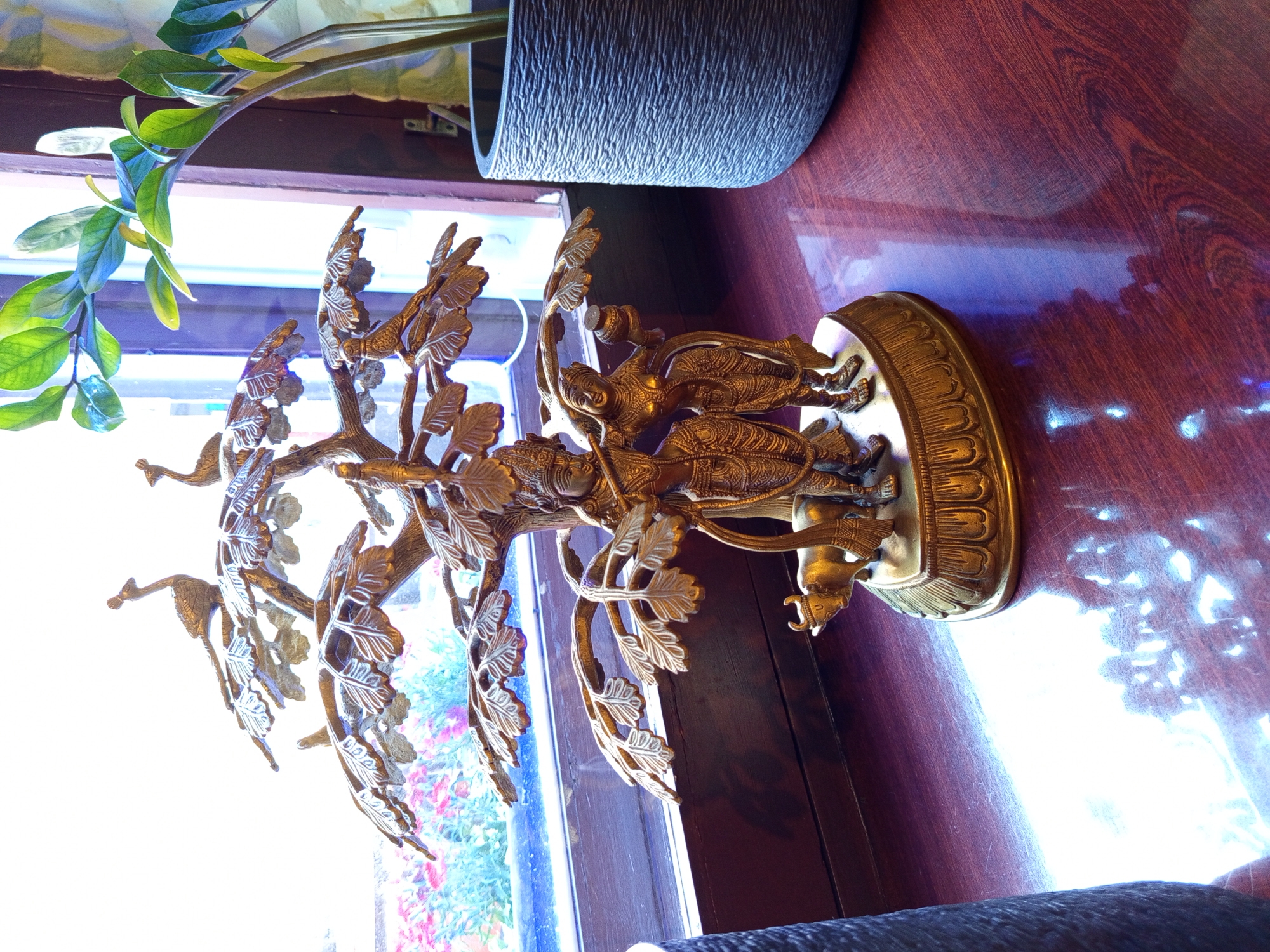 A brass statue depicting the Hindu deities Radha and Krishna is positioned on a polished wooden surface near a window in a building located in Saint Petersburg, Russia. The statue features Krishna playing a flute, with Radha standing beside him. Both figures are adorned in traditional Indian attire and are surrounded by a tree structure with intricate leaf designs. Perched on the branches of the tree are several small brass birds, resembling peacocks and sparrows. At the base of the statue, a small brass cow stands. The window behind the statue offers a blurred view of colorful flowers and sunlight. To the right of the statue, a grey textured planter holds a green plant. The wooden surface reflects the light and the objects placed upon it.