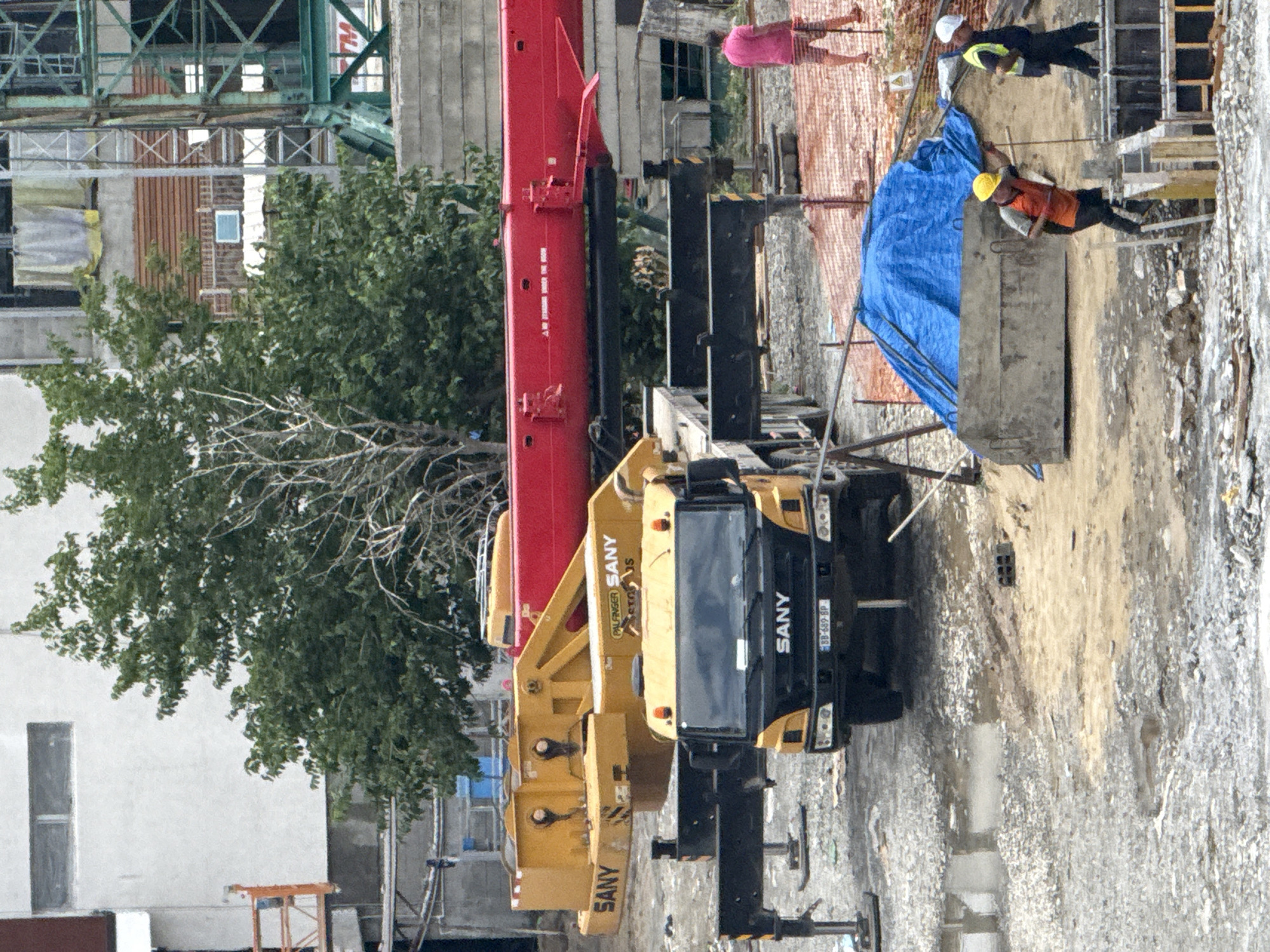 The image depicts a construction site, likely in Tbilisi, Georgia, featuring heavy machinery and workers. A large red crane boom extends horizontally across the frame, supported by yellow machinery labeled "SANY." A yellow SANY crane truck is positioned in the foreground, its stabilizers deployed. Two construction workers are visible: one in an orange vest and hard hat is ascending a ladder, and another in a yellow vest and hard hat is walking away from the camera. A person wearing a pink shirt and striped shorts stands in the background. The ground is uneven and appears to be a mix of gravel and mud, with puddles of water present. Construction materials, including concrete forms, are scattered around the site. A blue tarp covers an object in the middle ground. In the background, buildings and what appears to be a green construction crane are visible.