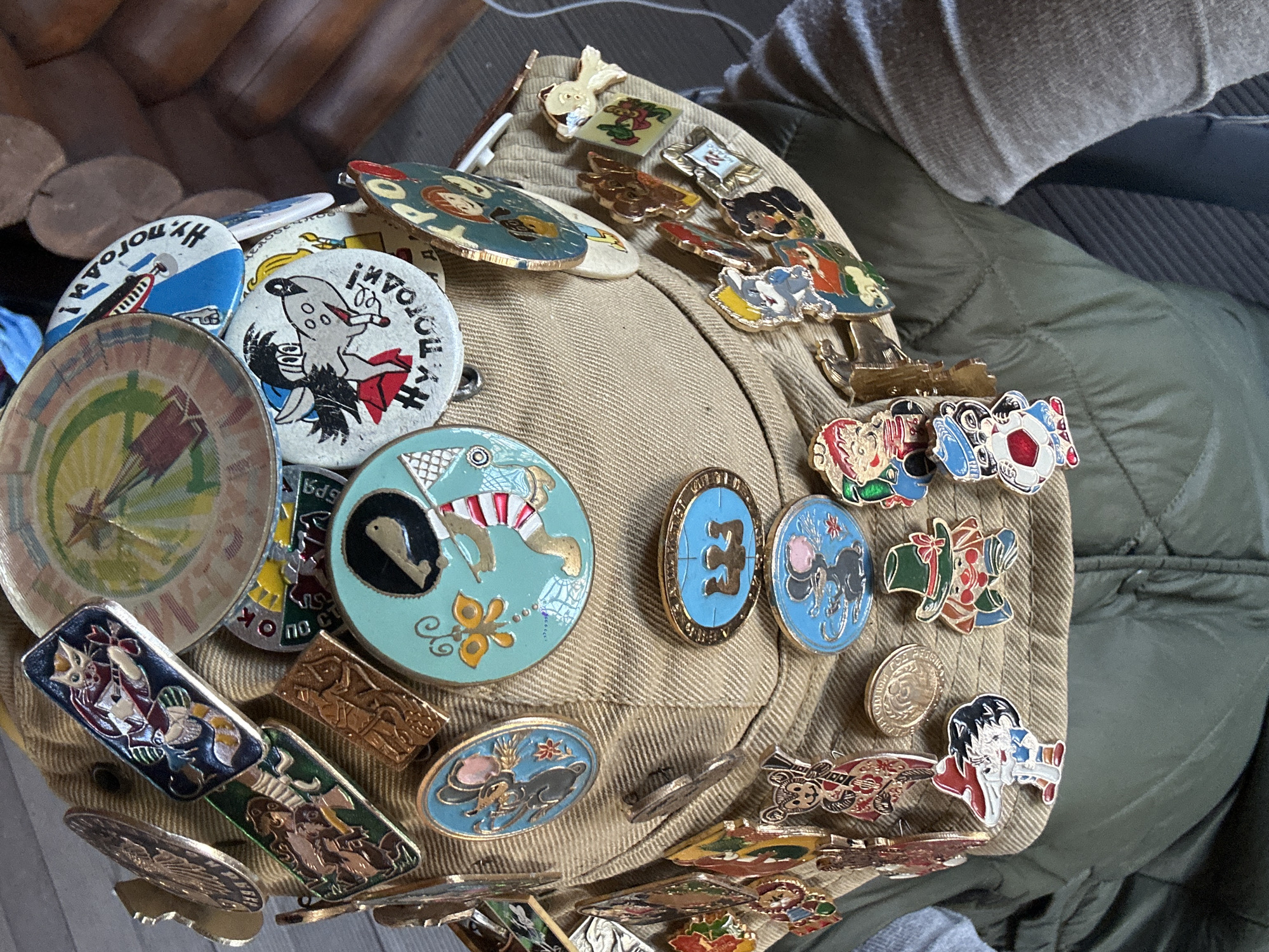 The image depicts a tan hat heavily adorned with pins, seemingly resting on a person's lap or near a garment. The pins appear to be a mix of Soviet-era and possibly more contemporary designs. Some of the pins have cartoon characters, geometric patterns, and images reminiscent of propaganda art. The background shows parts of a wooden object and a green quilted jacket. It cannot be confirmed this is in Nizhny Novgorod, Russia.