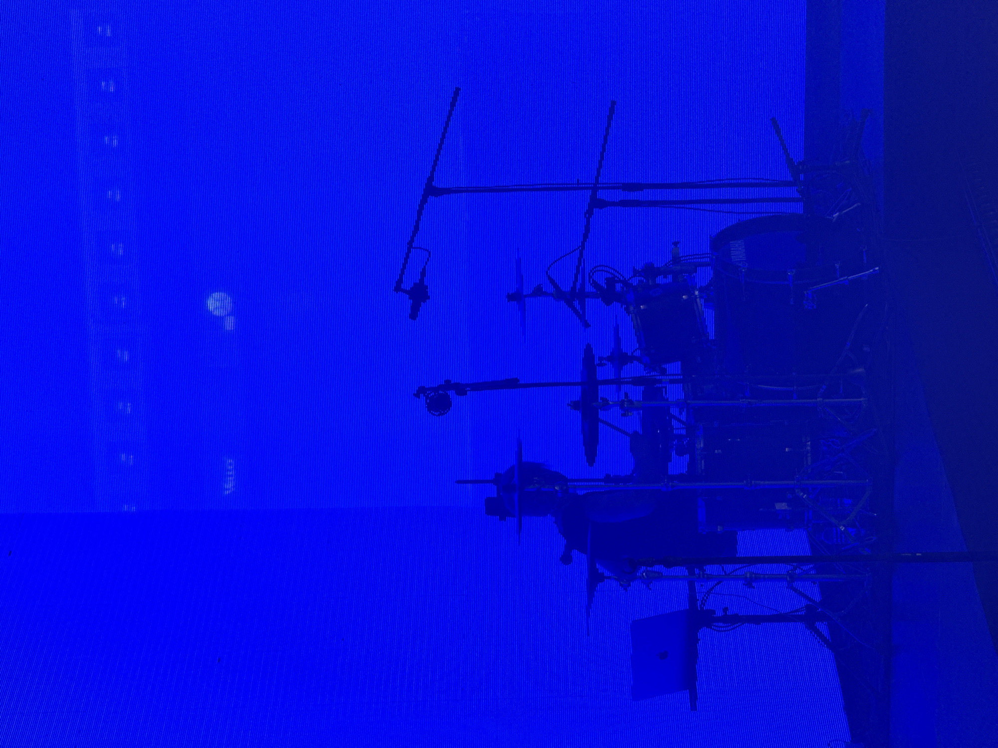 The image depicts a drum set illuminated by blue lighting. The drum set, which is a Yamaha brand, is situated on a stage, likely for a musical performance. The location of the drum set is not provided in the image.