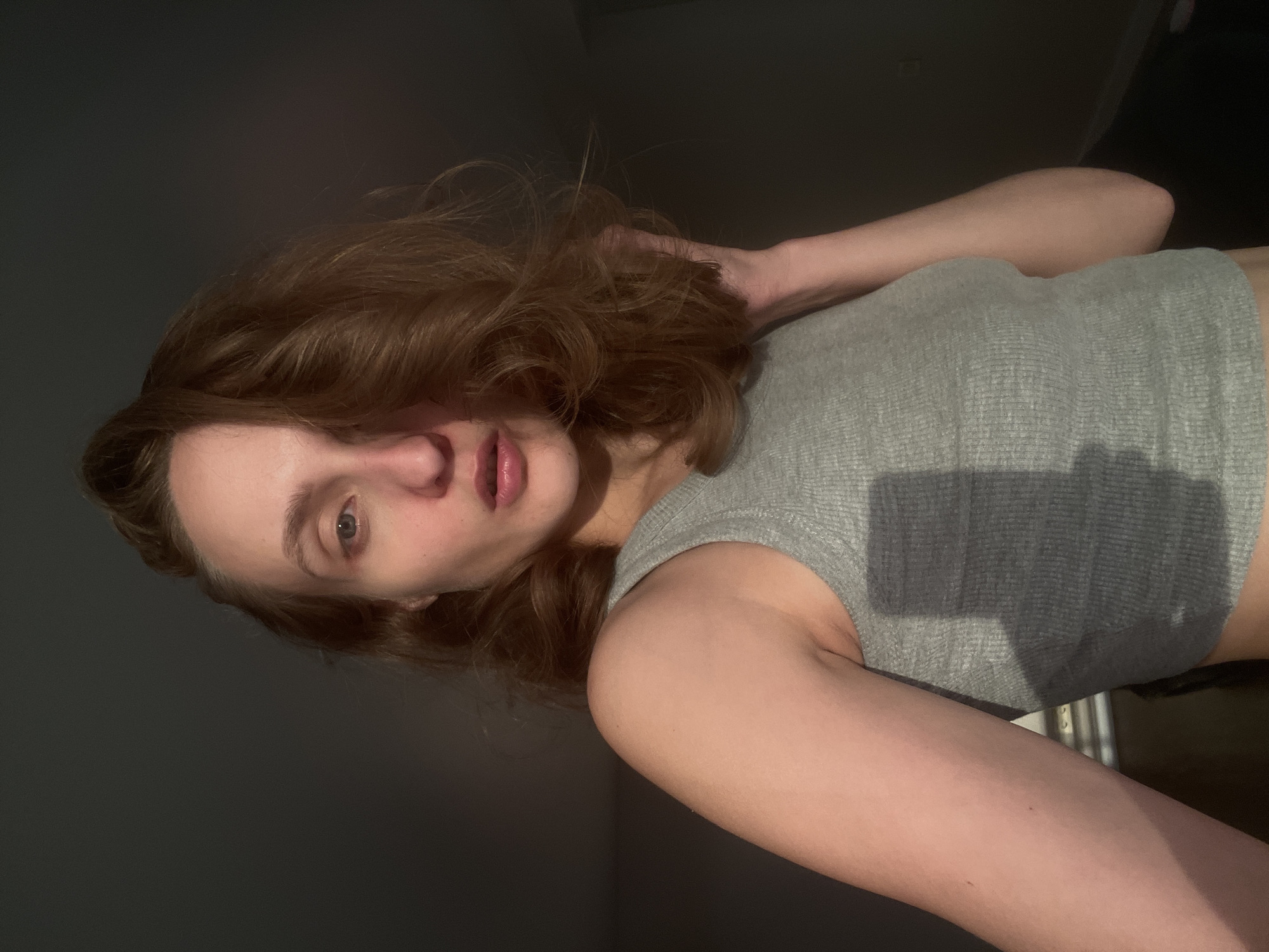 A young person with reddish-brown hair is taking a selfie in what appears to be a dimly lit room. The person is wearing a gray, ribbed tank top. Their hair is partially obscuring their face, and their left hand is resting on their head, behind their hair. The lighting suggests it is either early morning or late evening, with sunlight casting long shadows, possibly from a window. The background is dark and indistinct, with no clear architectural details to pinpoint the location further, aside from the provided context of Podshivalovo, Russia. The mood is casual and intimate, typical of a personal selfie. There is no visible text in the image.