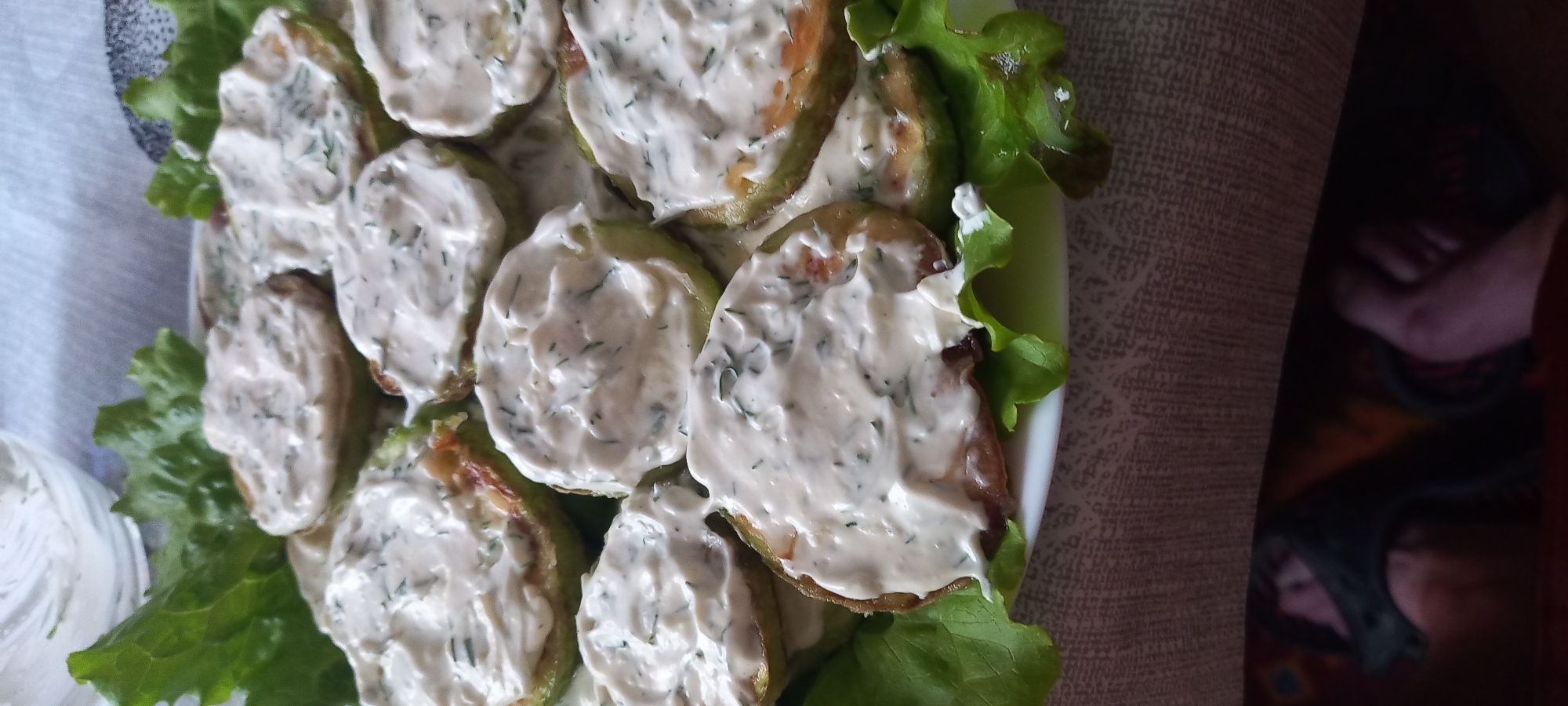 A close-up, high-angle image captures a white ceramic plate containing approximately ten to twelve round slices of pan-fried or baked zucchini, each generously topped with a creamy white sauce infused with chopped green dill. Fresh green lettuce leaves are interspersed among the vegetable slices on the plate. In the upper left background, a clear glass bowl holds an additional quantity of the same white dill sauce. The plate is situated on a light-colored, patterned tablecloth. The lower portion of the image reveals, partially obscured by shadow and red fabric, a bare human foot and a dark object, possibly footwear, indicating the presence of an individual in the immediate vicinity. No specific geographical markers are present to identify the location as Kryvyi Rih, Ukraine; the scene is consistent with a domestic setting.