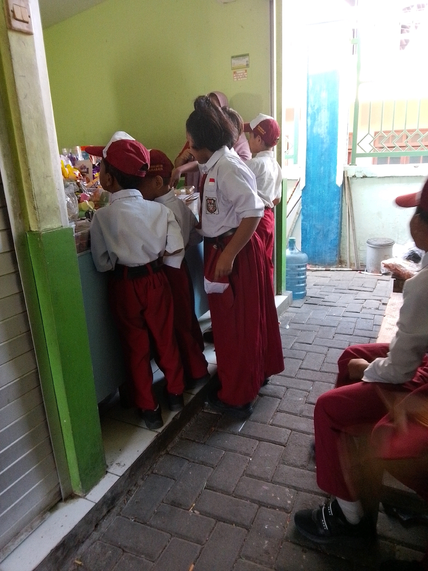 In Surabaya, Indonesia, near a school, a group of children in white shirts and red pants or skirts stand near a food stall. The stall has various food items and drinks on display. The children appear to be selecting or purchasing food. The environment is outdoors, with a paved walkway and some greenery visible. The children are wearing school uniforms, suggesting this is during school hours. One child in the foreground is holding a snack bag. The overall scene depicts a typical school day in Surabaya, with children enjoying a snack break.