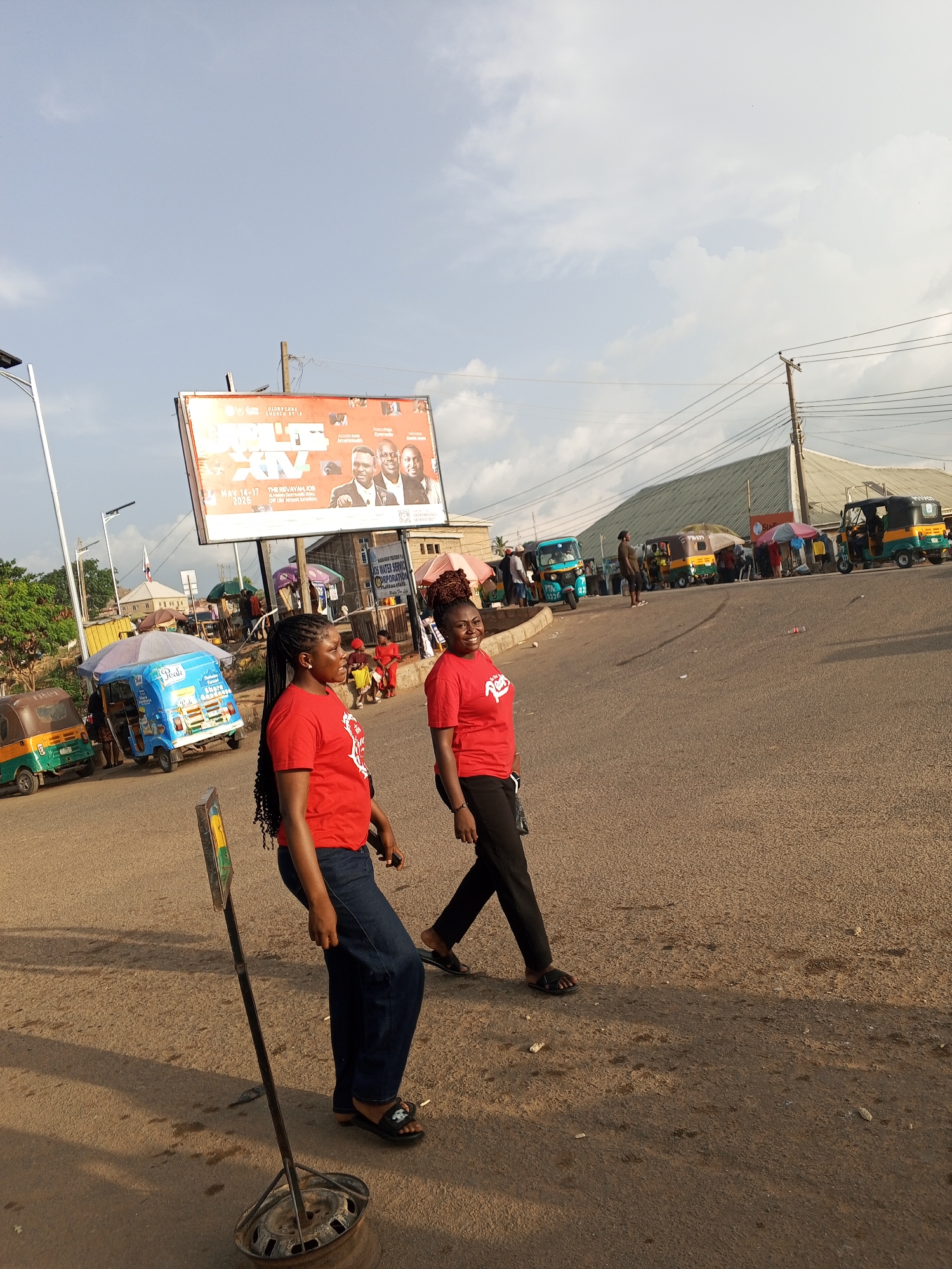 The scene captures an urban environment in Gwong, Nigeria, featuring two women dressed in matching red t-shirts walking along a paved road. A large billboard advertising a "Revayah" event dated May 14-17, 2025, stands prominently in the background. Several auto rickshaws (tuk-tuks) are parked along the roadside, indicating a local mode of transportation. Additionally, street vendors or stalls can be seen with people gathered around. In the foreground, a damaged road sign attached to a discarded tire rim lies beside the road.
