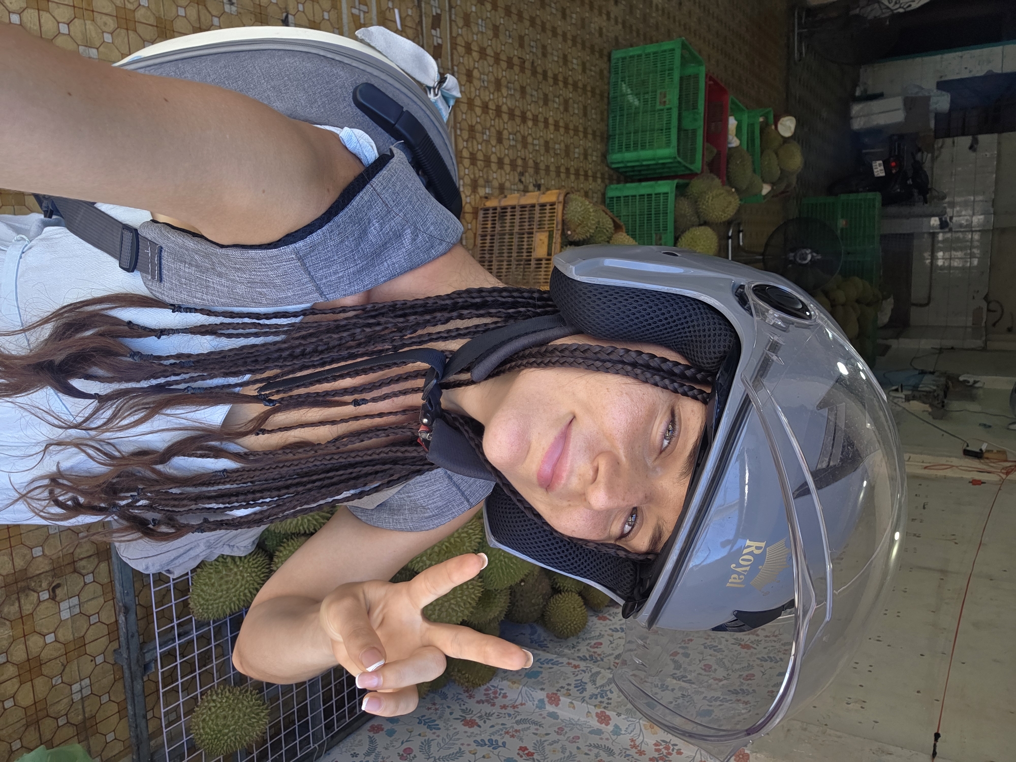 A woman is taking a selfie in an outdoor market in Phan Thiết, Vietnam. She is wearing a gray backpack and a motorcycle helmet. Her dark hair is styled in many braids that hang down her chest. She is smiling and making a peace sign with her right hand. In the background, there are many durians for sale, some in green crates and others in a metal cage. The market appears to be open-air, with a tiled floor and a corrugated metal roof visible in the background. The time of day appears to be daytime, and the weather seems to be warm and sunny, typical for Vietnam. The woman's expression conveys happiness and excitement. The word "Royal" is visible on her helmet visor.