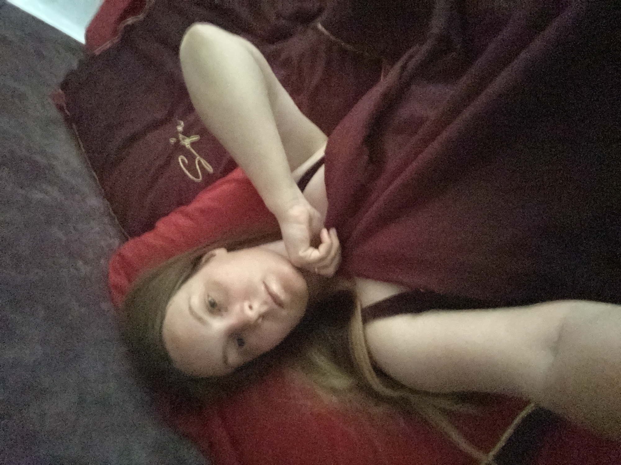 A woman lies on a bed covered with maroon and red bedding. The bed appears to be on a grey surface, likely a couch or another section of the bed. She is partially covered by the maroon bedding, with one arm raised and her hand near her face. She is wearing a dark top or bra. Her head rests on a red pillow. Her hair is light brown and shoulder-length. The image quality is slightly grainy. The location cannot be determined to be in Noviy, Russia as there are no details to confirm it.