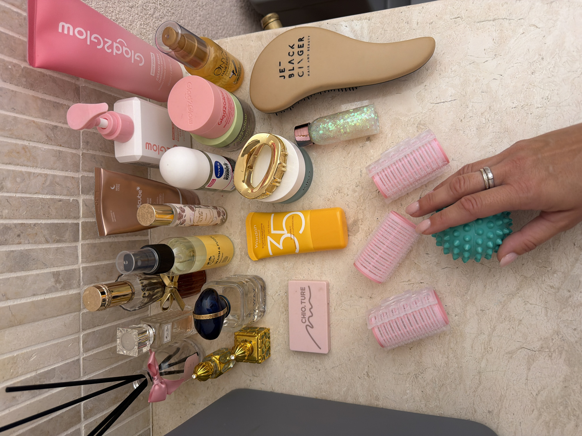 The image displays an assortment of personal care and beauty products arranged on a speckled, light-colored countertop, likely in a bathroom setting, as indicated by the tiled backsplash. Several perfume bottles, some with decorative caps and ribbons, are present. There is a reed diffuser with black reeds. A bright yellow sunscreen tube with the brand "Wardah" is visible, alongside a roll-on deodorant, a pink pump bottle, and various jars and small bottles, some containing lotions or serums, including a tube with "Glad2Glow" branding. A beige hairbrush with black lettering that reads "JE BLACK GINGER HAIR AND BEAUTY" is positioned to the right. Scattered among the larger items are smaller accessories: pink hair rollers, a turquoise spiky massage ball, and a small decorative bottle filled with glitter. A person's hand, adorned with rings, is partially visible in the lower right portion of the frame, interacting with the massage ball. The overall scene suggests a collection of items used for personal grooming and beauty routines. The location is not discernible from the image.