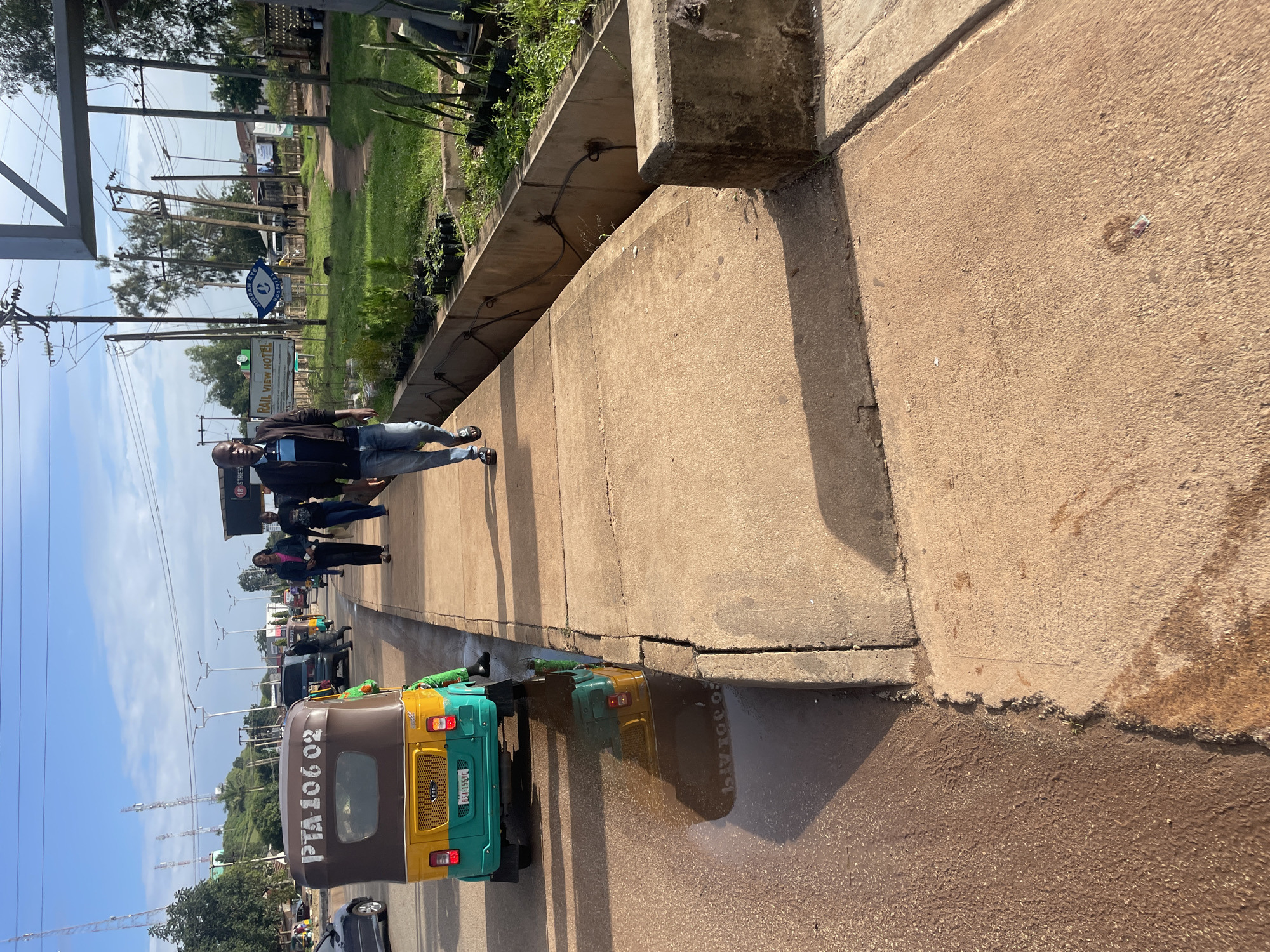The image captures a street scene in Jos, Nigeria, likely during the daytime with clear skies. In the foreground, a yellow and teal colored auto-rickshaw with the license plate "RSA 155JC" and the inscription "PTA 106 02" is parked at the edge of a road. The road surface is wet, with a large puddle reflecting the auto-rickshaw. A sidewalk made of concrete runs alongside the road. A man in a dark jacket, blue jeans, and black sandals is walking on the sidewalk away from the viewer. Further down the sidewalk, two women are walking in the same direction, one in a pink top and black pants, and the other in a dark top and jeans. In the background, there are more vehicles, trees, utility poles with wires, and buildings, including one with a sign that reads "RAIL VIEW HOTEL". The overall scene depicts a typical urban environment in Jos.