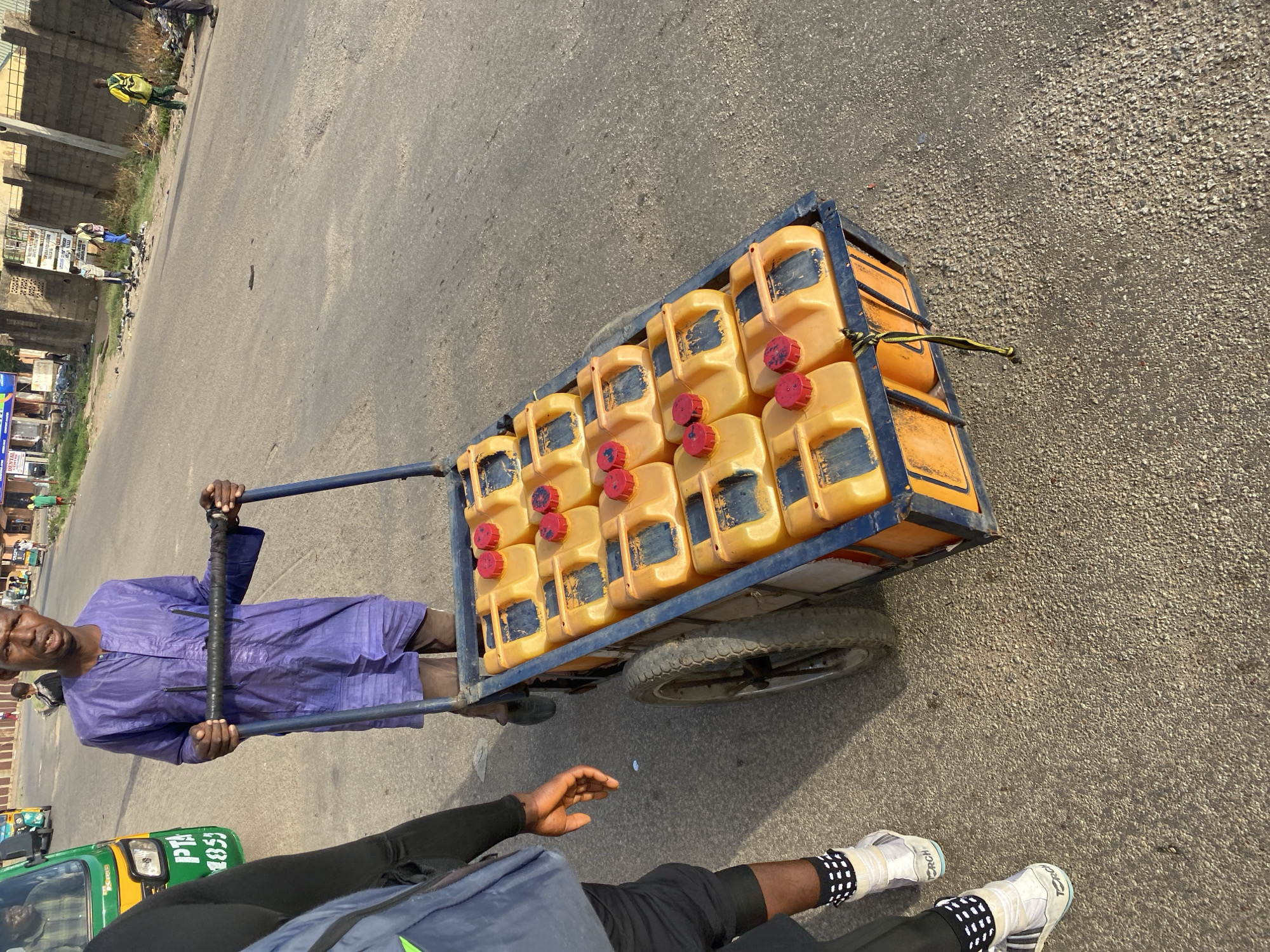 A man wearing a blue robe pushes a blue metal cart laden with approximately 20 yellow plastic jerrycans, each with a red cap, down an asphalt road in Jos, Nigeria. Another person, identifiable by legs and feet clad in black pants, white shoes, and dotted socks, stands nearby. In the background, there is a street with buildings and a vehicle visible.