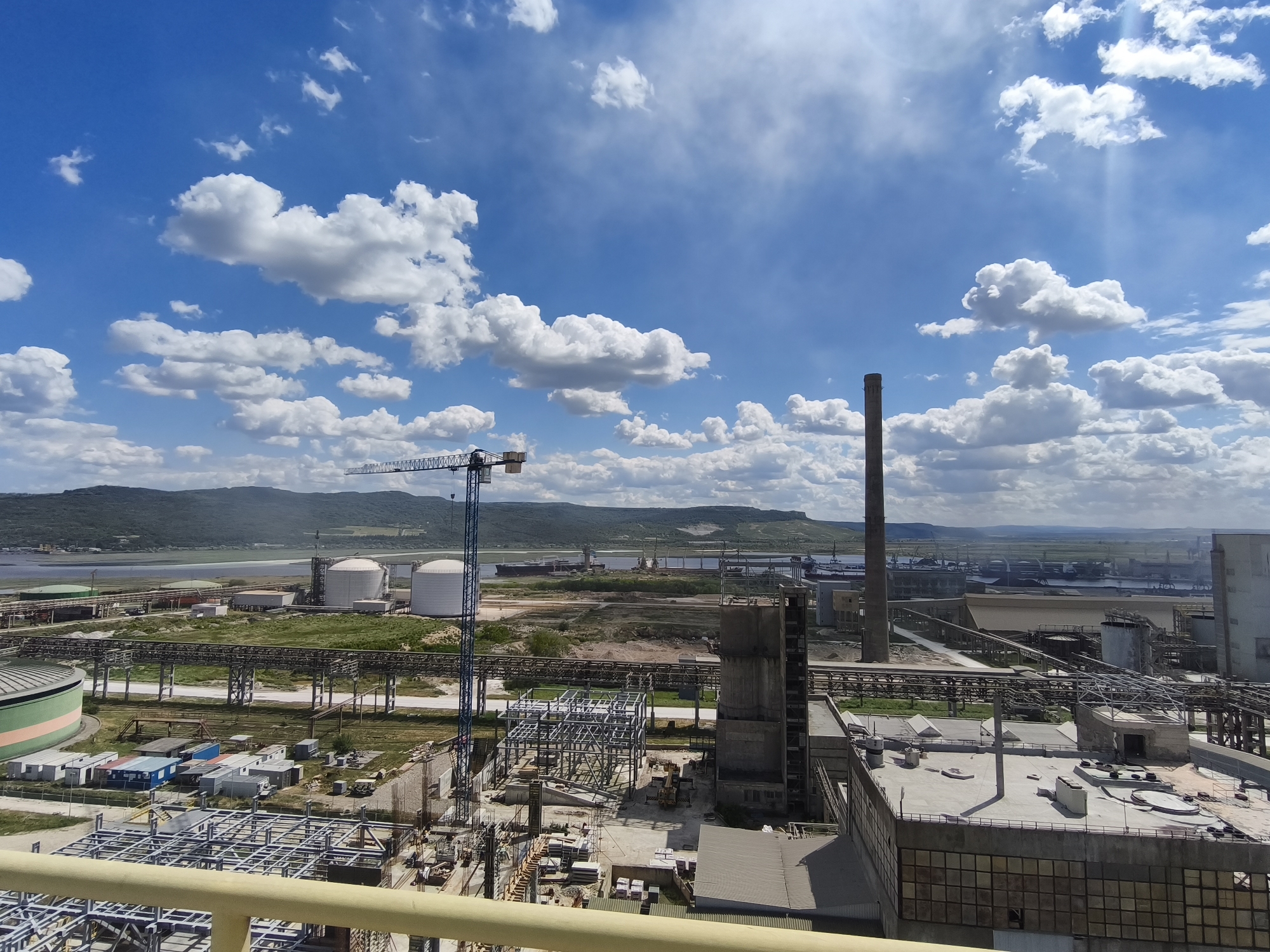 The image depicts an industrial site, potentially a factory or refinery, located near a body of water. A tall smokestack and a construction crane dominate the skyline against a backdrop of blue sky with scattered white clouds. Several large storage tanks are visible in the midground, connected by a network of pipes and structures. In the foreground, there's ongoing construction with exposed steel framework and various construction materials. Older buildings with visible wear and tear are also present, suggesting a mix of new development and established infrastructure. The location is near hills and water, with ships visible in the distance.