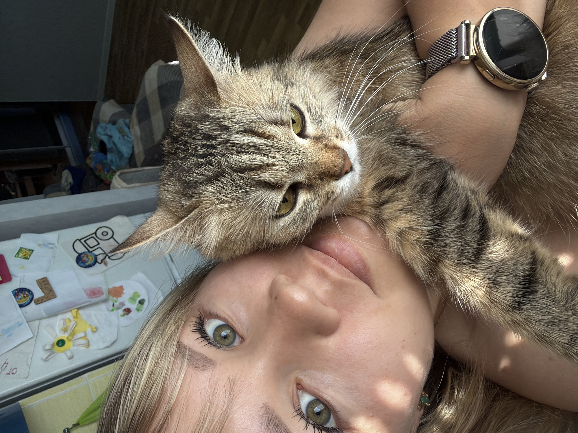 A woman with blonde hair and green eyes is holding a tabby cat close to her face. She is wearing a smartwatch with a metal band on her left wrist. The background includes a window, a couch, and a table with various objects on it. The location is in the city of Chaikovsky, Russia. The woman and the cat are interacting in a close, affectionate manner.