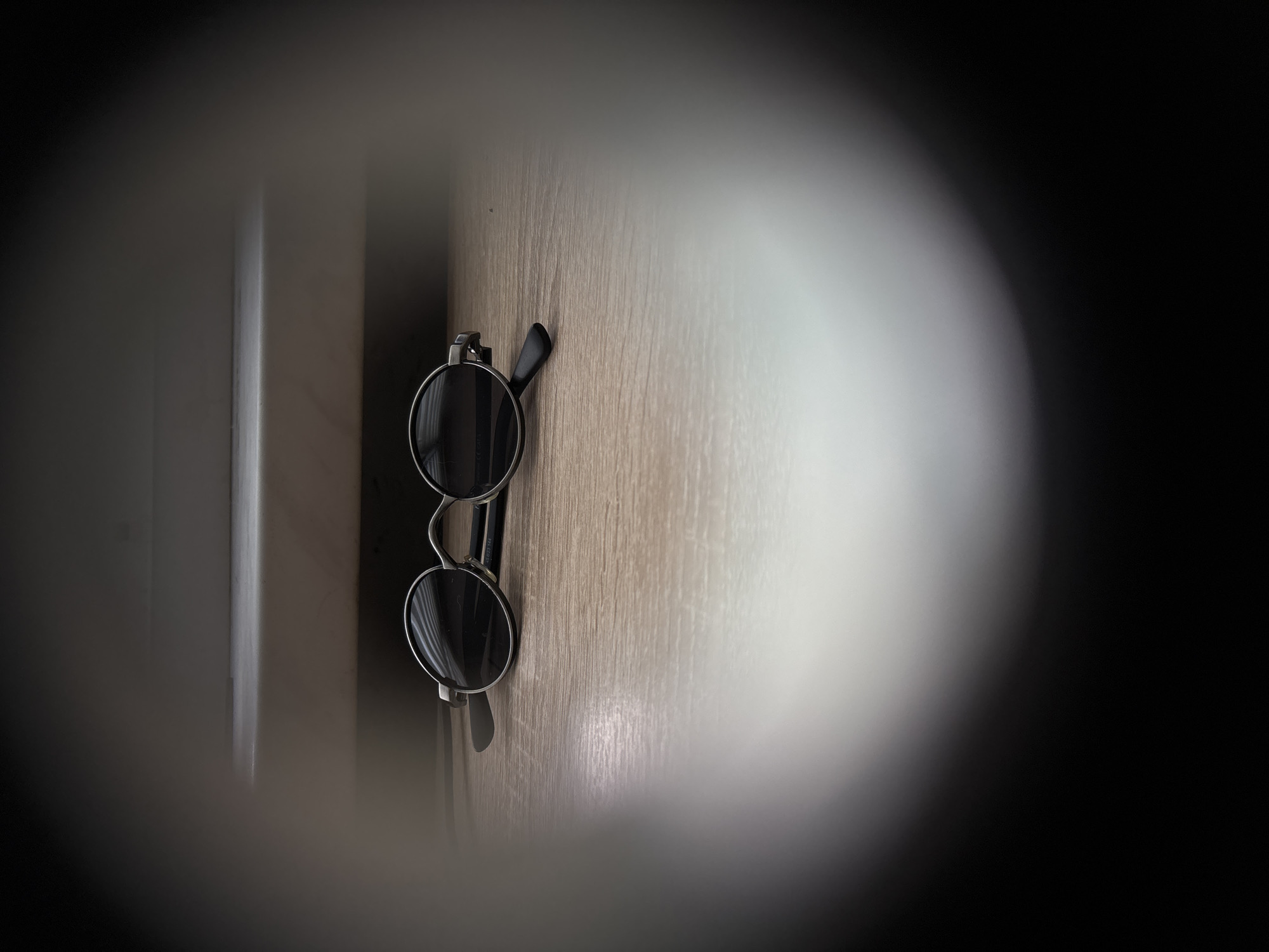 A pair of round, black-rimmed sunglasses with dark lenses rests on a wooden surface. The sunglasses are positioned horizontally, with the temples folded inwards. The wooden surface appears to be a table or shelf with a visible grain pattern. In the background, a slightly lighter-colored horizontal surface, possibly a shelf or ledge, is present. The image is framed by a soft, circular vignette, blurring the edges and creating a tunnel-like effect. The lighting is dim, with a brighter area towards the bottom right corner of the frame. There is no indication of the location being in Saint Petersburg, Russia.