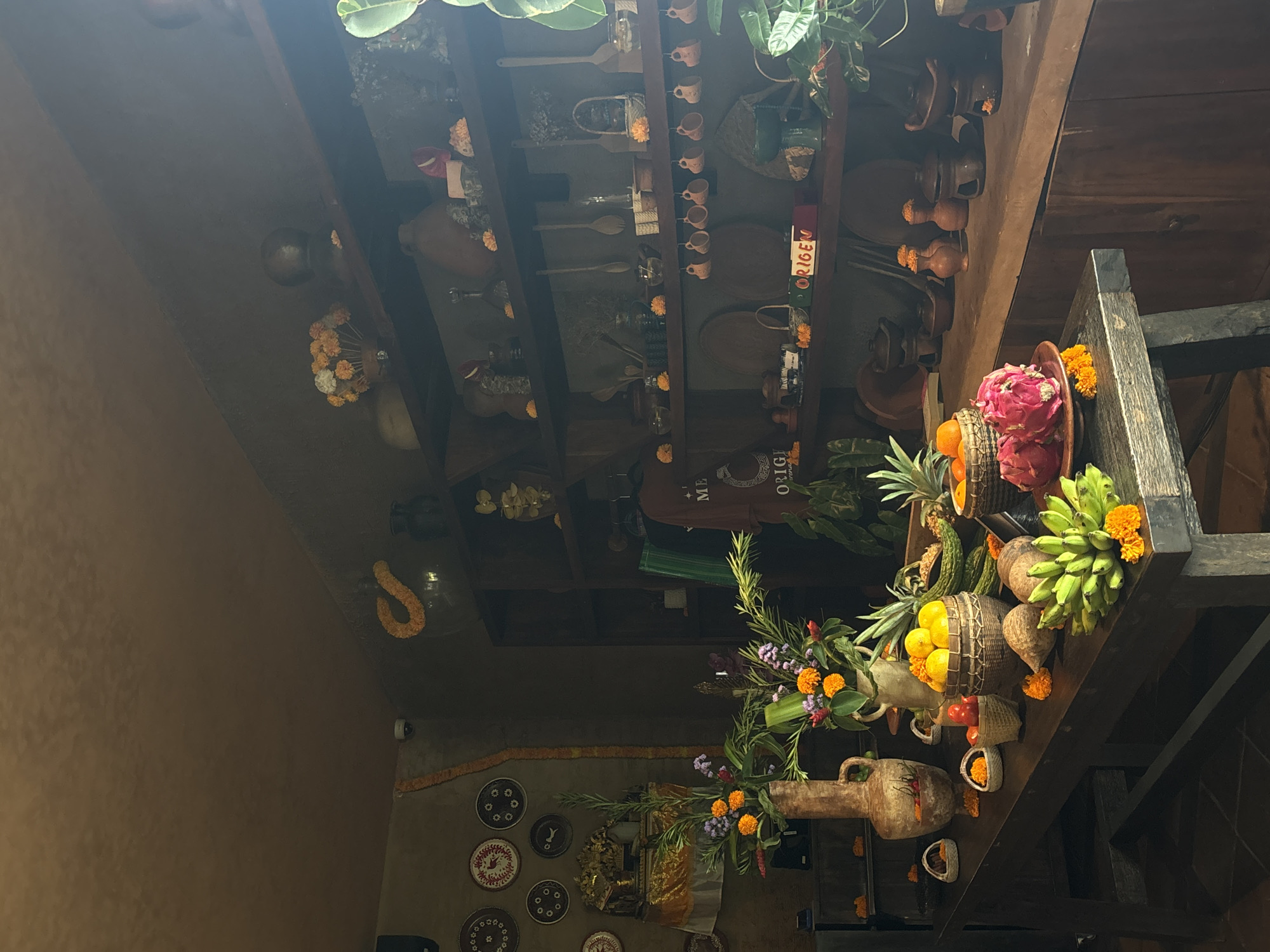 Captured indoors, the image showcases a shelf laden with fruits and flowers, positioned near a wall adorned with decorative plates. This tableau is found in Mengwi, Indonesia. The wooden shelf is multi-tiered and dark-stained. Atop it, one finds baskets filled with assorted fruits such as oranges, dragon fruit, and green bananas. These fruits are punctuated by orange flowers.
Below the fruit display is a vase brimming with floral arrangements, predominately of purple and orange hues. The wall behind the shelf is textured and brown, decorated with a string of orange flowers and multiple decorative plates. The plates have various designs, adding a cultural element to the scene.