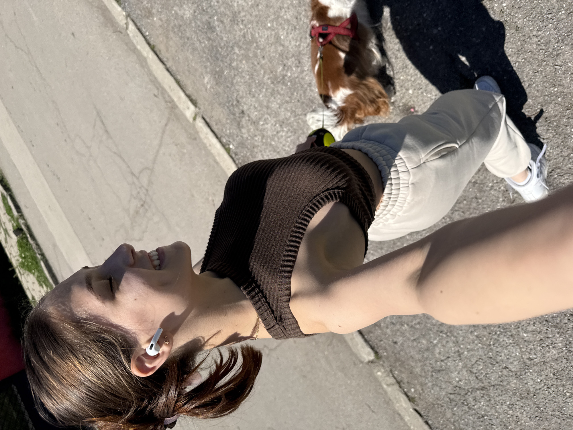 A young woman is taking a selfie while walking a dog on an urban street in Sofia, Bulgaria. The woman is fair-skinned, has brown hair pulled back, and is wearing a brown ribbed tank top, light gray sweatpants, and white sneakers. She has white earbuds in her ears. She is smiling and facing towards the sun. Next to her is a small dog with brown, black, and white fur, wearing a red harness. The dog is on a leash. They are walking on a paved street with a curb on one side. Their shadows are visible on the pavement. The photo is taken from a high angle, likely by the woman herself.
