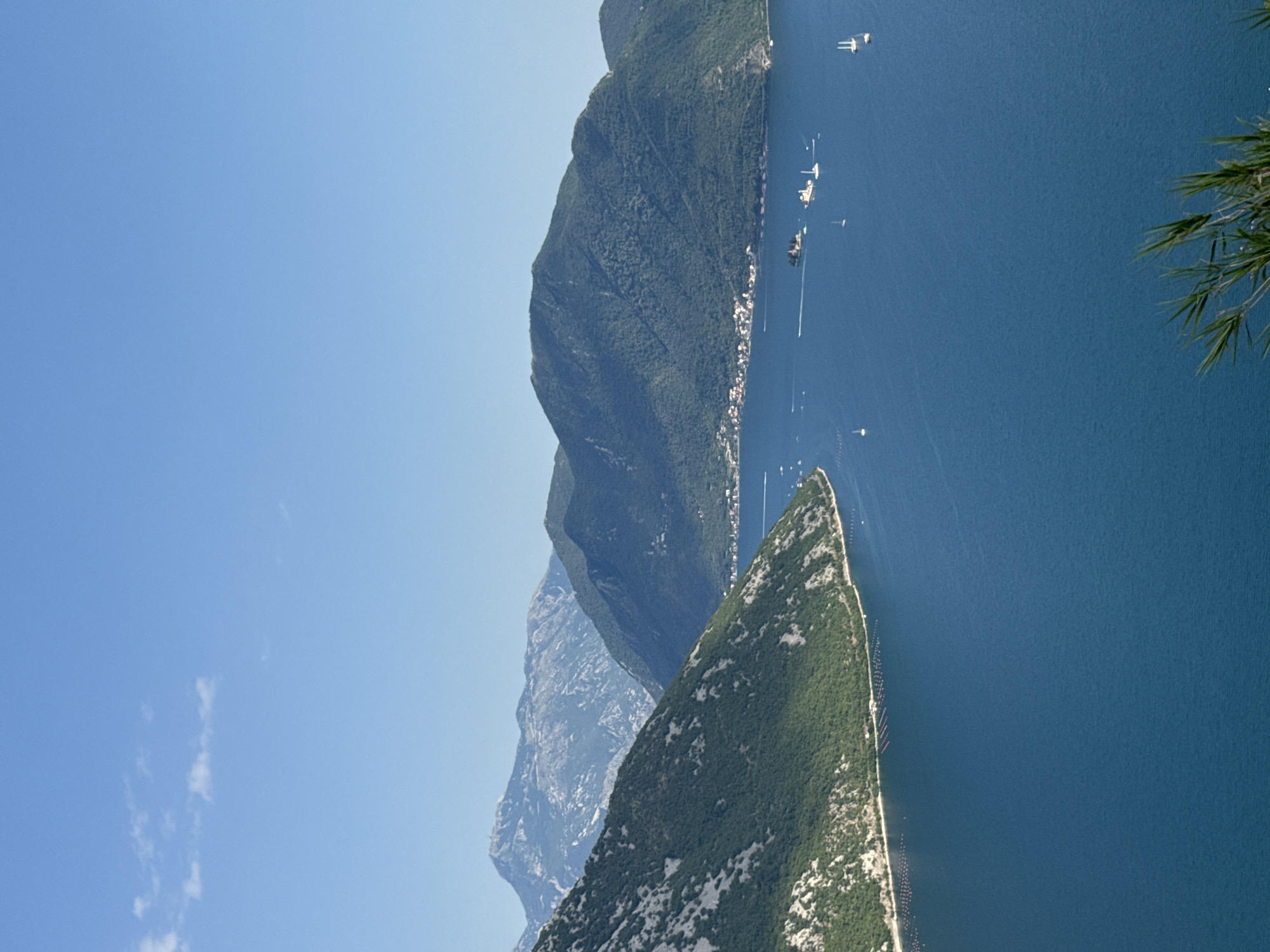 The image captures a scenic view of the Bay of Kotor in Montenegro, with a focus on the town of Štrp. In the foreground, lush green mountains covered in trees slope down towards the vibrant blue water. The bay is dotted with several boats, including sailboats and what appear to be ferries, traversing the calm waters. In the mid-ground, a small island, likely Sveti Đorđe or Gospa od Škrpjela, is visible with buildings and a church. A coastal town with red-tiled roofs, characteristic of Montenegro, is nestled along the shoreline. In the distance, more mountains rise, some with rugged, rocky peaks, creating a dramatic backdrop. The sky is a clear, bright blue with a few wispy clouds. The overall impression is one of a serene and picturesque coastal landscape.
