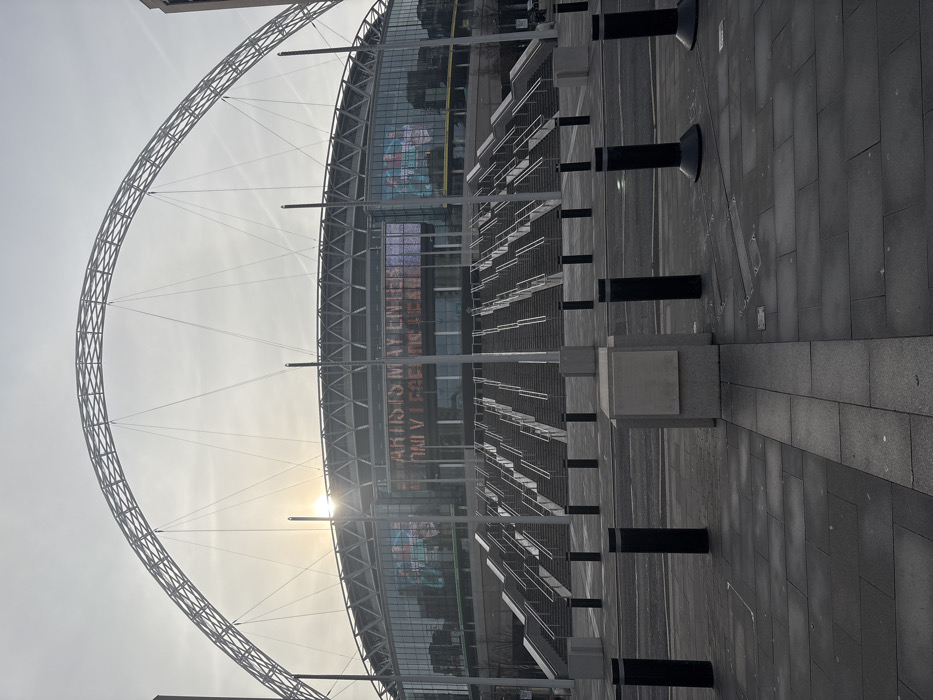 The image captures the exterior of Wembley Stadium in Wembley, United Kingdom, under bright, slightly hazy conditions, likely during early morning or late afternoon due to the low sun. The scene is dominated by the stadium's distinctive white arch, which spans gracefully across the sky above the main structure. The sun's glare is prominent behind the stadium's facade, particularly to the left of its central point, indicating its low position.
The stadium building features a modern design with large glass panels and a visible structural framework. On one of its exterior screens, faint white text is displayed. Partially legible phrases include "ARTISTS MAY ENTER" and fragments such as "IN REAL" and "UNIVERSE," possibly part of an advertisement or a general message.
In the foreground, a wide, paved plaza or concourse extends towards the stadium. This area is completely devoid of people, suggesting it is not an event day or that the image was taken during off-peak hours. Several black, cylindrical bollards are systematically arranged along the paved walkway, interspersed with square concrete plinths. Further back, metal barriers or queueing structures are visible on the steps leading up to the stadium entrance. The overall impression is one of architectural grandeur and scale, captured in a moment of quiet anticipation or dormancy.