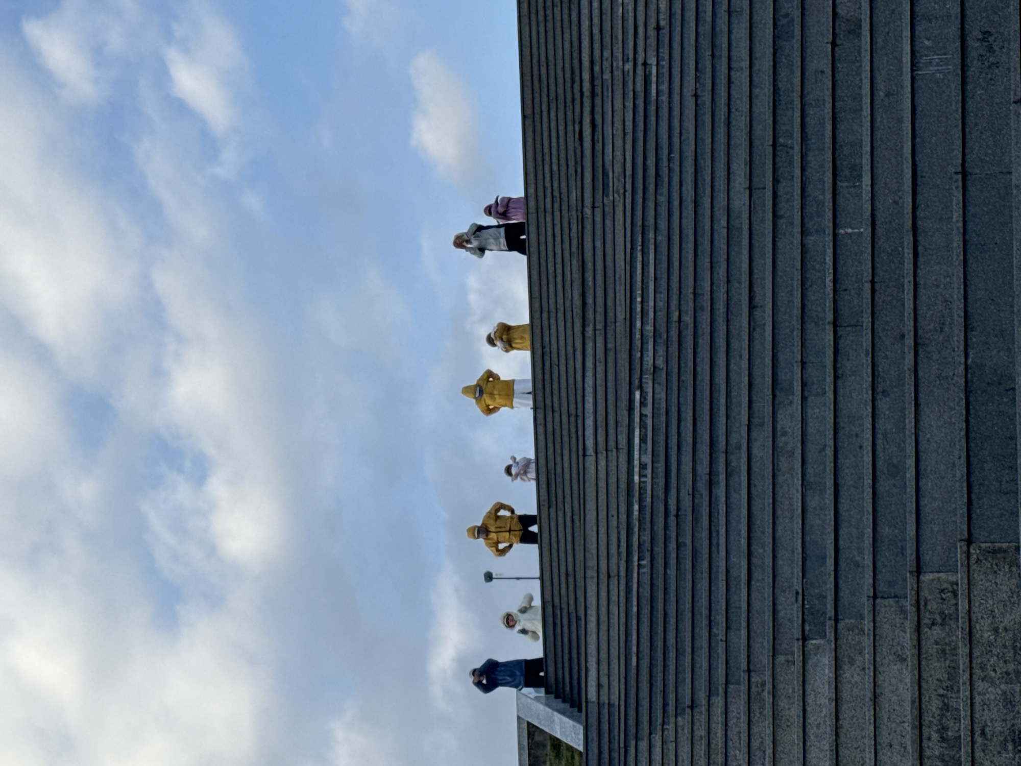 A low-angle photograph shows seven people standing on a concrete staircase in front of a cloudy sky. The staircase has a vertical, striped texture created by the individual steps. The people, wearing hooded jackets of various colors (blue, white, yellow, pink, and grey), stand in a line, facing the viewer. Some have their arms raised, as though taking photos. The location is likely Da Lat, Vietnam.