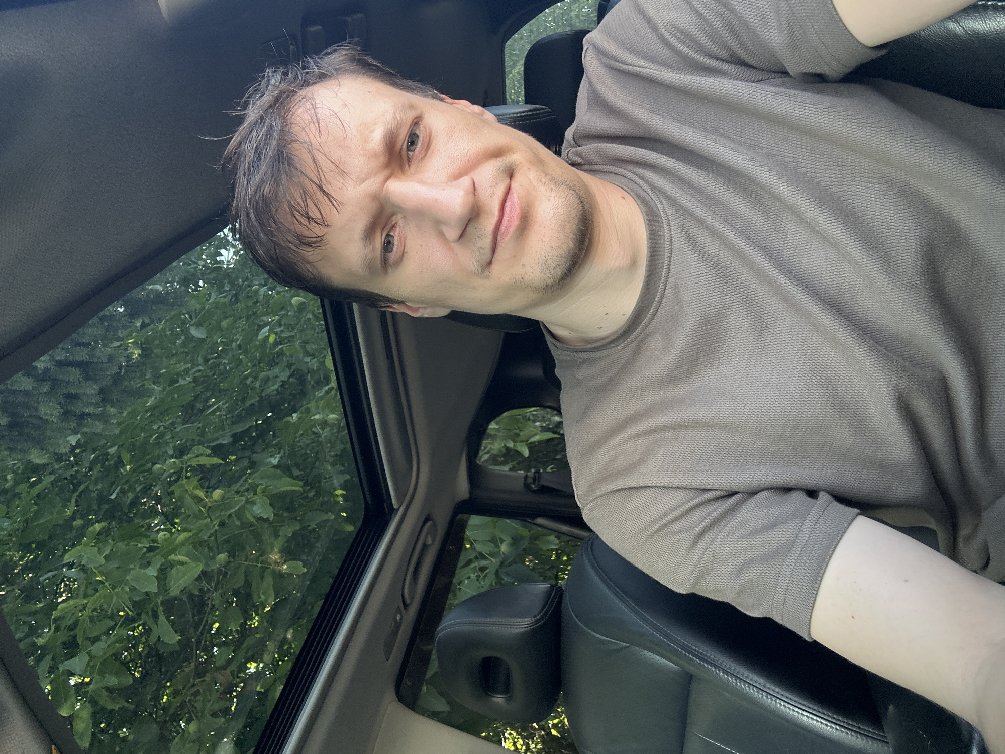 A man is sitting in the driver's seat of a car, looking at the camera. He has short, brown hair and is wearing a grey t-shirt. The car has a sunroof, which is open, revealing trees and foliage above. The interior of the car is visible, including the black leather seats and dashboard. The lighting suggests it is daytime. The location is Shatava, Ukraine.