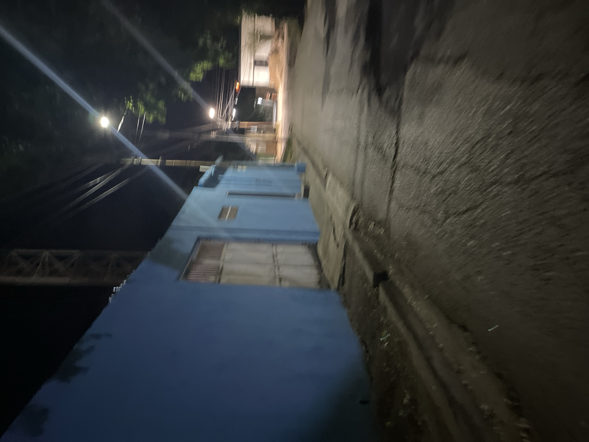 A night-time street scene in Santa Ana, Venezuela, features a blue stucco building with large, closed metal double doors on the left. A narrow concrete gutter runs along the base of the building, filled with debris. The street itself is paved, appearing wet and reflective in places, with visible cracks and patches. Overhead, power lines crisscross the dark sky, illuminated by the bright beams of streetlights. Further down the street, other buildings are visible, including one with white walls and dark-shuttered windows. Trees with dense foliage are present, casting shadows and contributing to the overall darkness of the scene. The overall impression is one of a quiet, sparsely populated urban environment after dark.