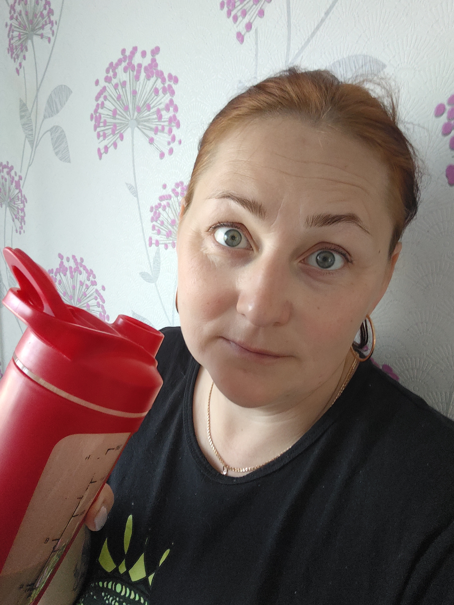 A woman with reddish-brown hair and green eyes is holding a red protein shaker bottle. She is wearing a black t-shirt and a gold necklace and earrings. The background is a wall with a floral pattern consisting of white flowers with pink spherical clusters on gray stems. Based on prior information, the location is Markovskii, Russia.