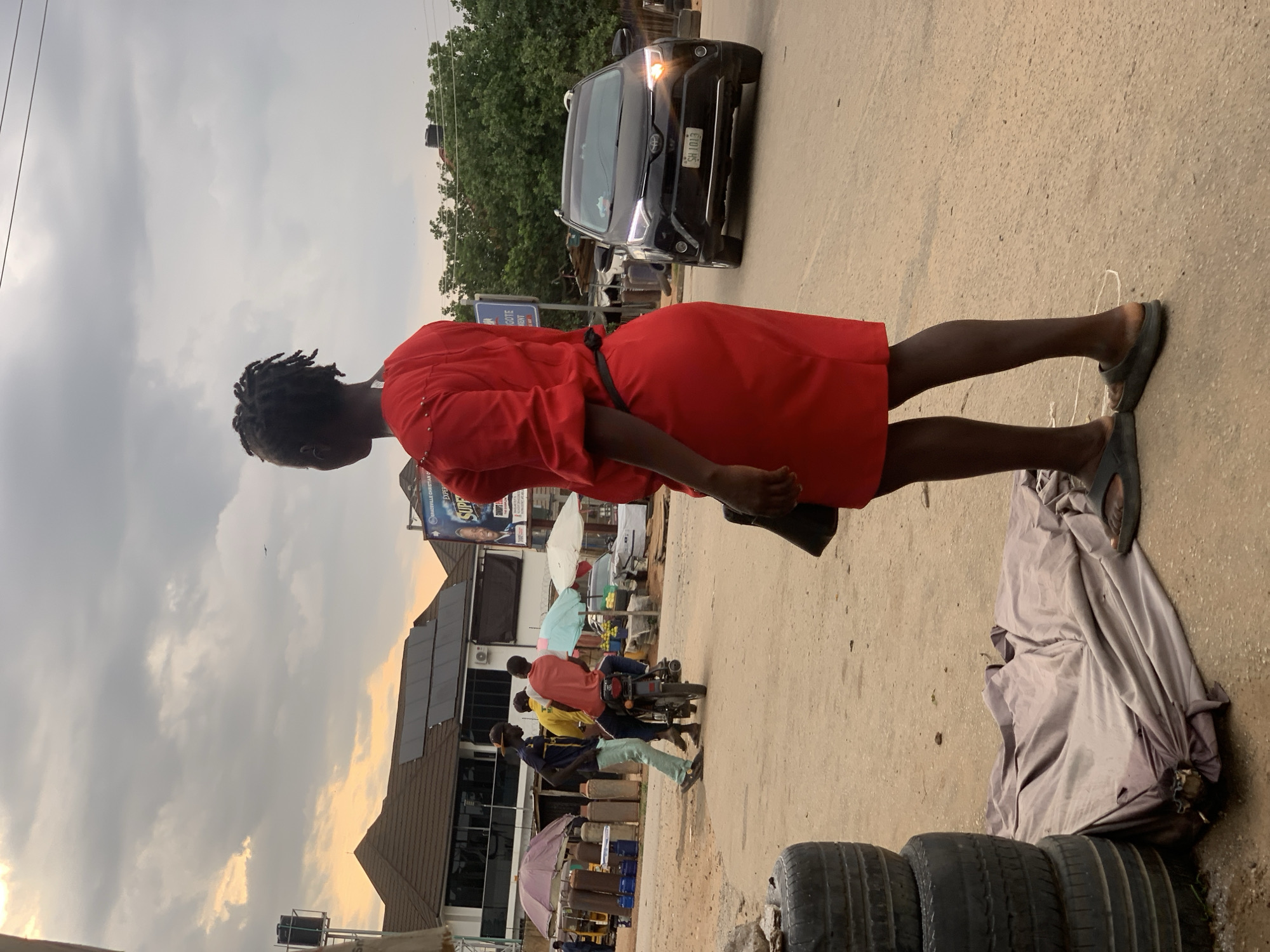 In Abuja, Nigeria, a woman in a red dress stands on a dusty road, facing away from the camera. Her hair is styled in braids. To her left, three men are gathered around a motorcycle, one on it and two standing. Behind them, various market stalls are visible with umbrellas, gas cylinders, and produce. A black Toyota SUV is driving on the road to her right. The sky above is cloudy, with hints of an approaching sunset. The scene appears to be a busy street in a commercial area.