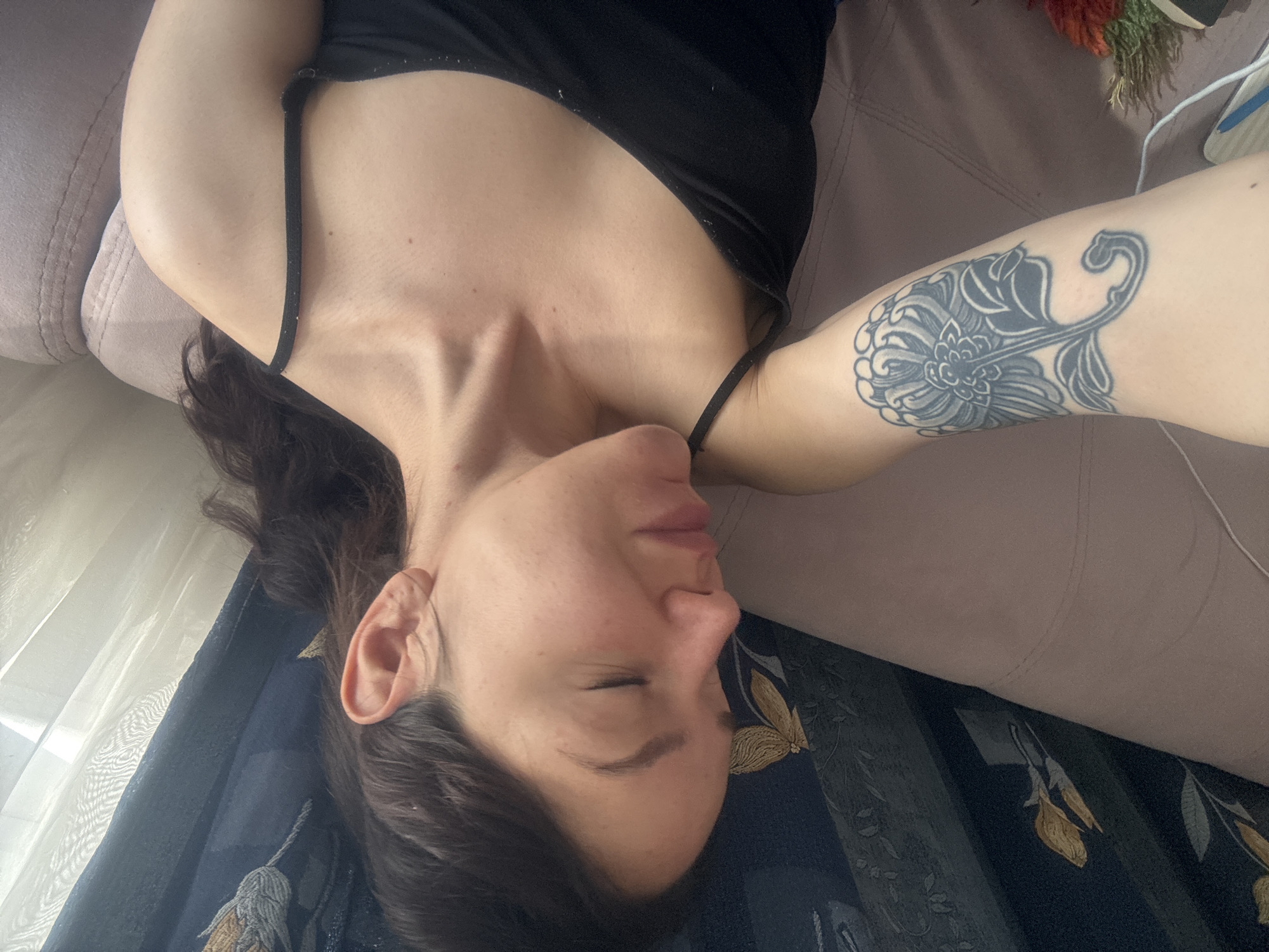 A woman with dark hair is lying on a couch in an indoor setting, possibly in Burgas, Bulgaria. She is wearing a black tank top. Her eyes are closed and she has a tattoo on her left forearm. A floral patterned fabric drapes near her head. The couch is a muted pink color. Some dried flowers and a white wire are visible in the background.