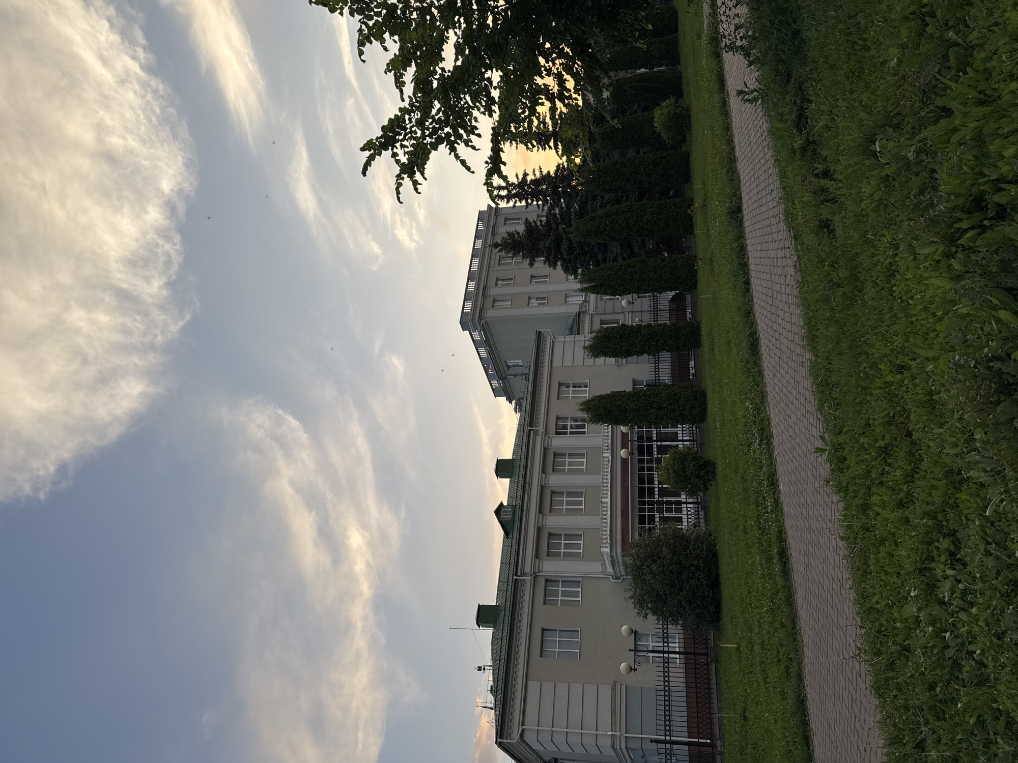 This is a photograph of a building in Izhevsk, Russia, taken during what appears to be sunset or early evening. The building in the foreground is a light-colored, multi-story structure with a green roof and numerous windows. A black metal fence runs along its perimeter. In front of the building is a paved walkway and a well-maintained grassy area. Behind the foreground building, another taller, light-colored building is visible, with a gray facade and a row of decorative elements along its roofline. A line of tall, conical evergreen trees stands between the two buildings, and a larger deciduous tree with lush green foliage is prominent on the right side of the frame. The sky above is a pale blue with wispy white clouds, tinged with the warm colors of sunset. Several small birds are in flight against the sky. The overall impression is of a calm, institutional or administrative setting.
