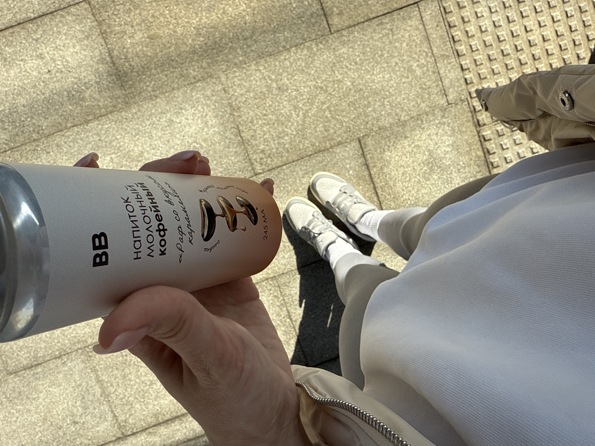 The image shows a person standing on a concrete tiled surface in an urban environment. The person is holding a can of a beverage, "BB молочный кофейный" (milk coffee). The can is predominantly white with brown accents and Russian text. The person is wearing white sneakers, white socks, and light-colored clothing, including a jacket with a visible zipper. The photo is taken from a high-angle perspective.