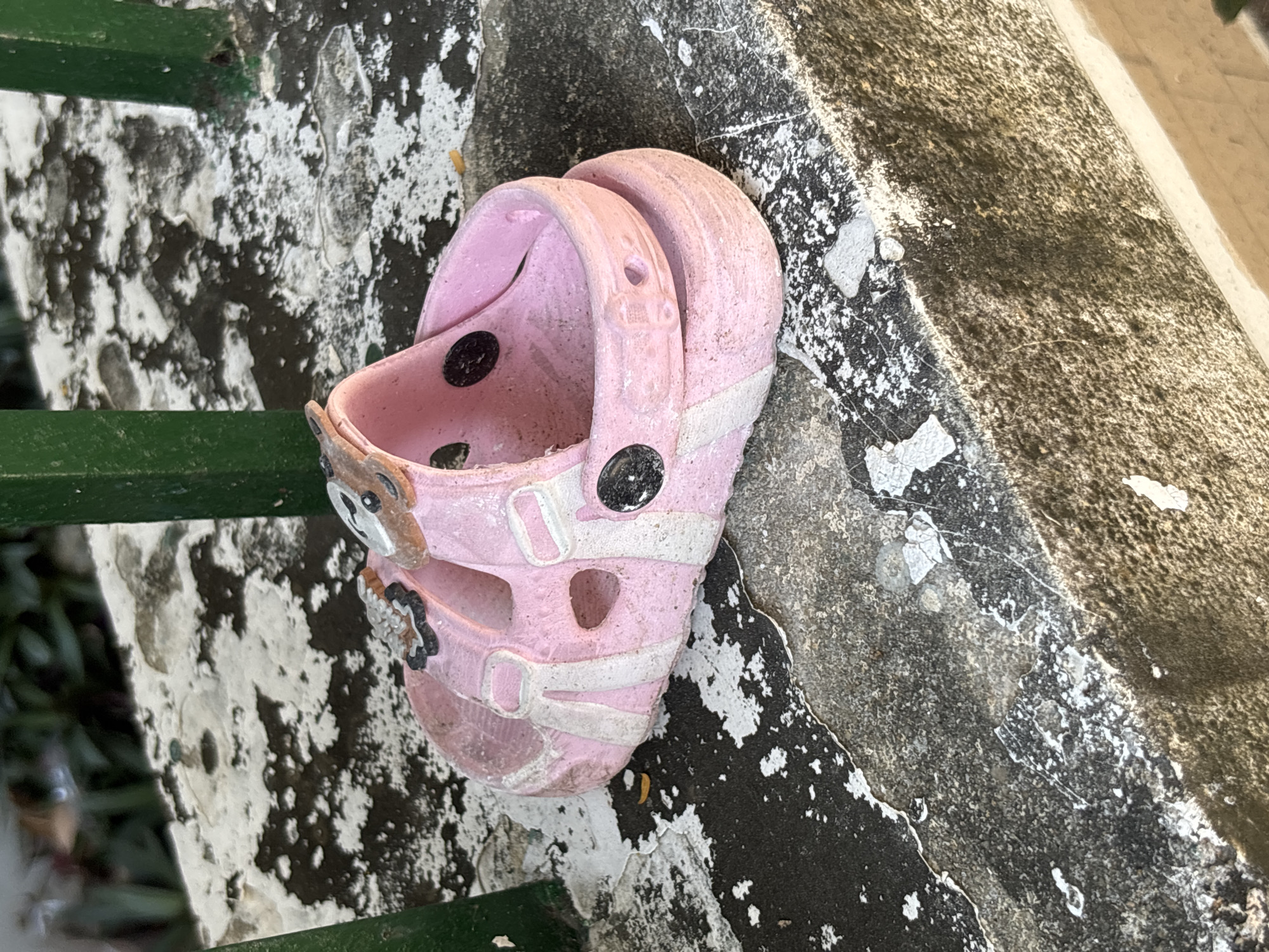 A single, pink child's sandal, likely a clog-style shoe with a strap and decorative embellishments, rests on a weathered, grey concrete ledge. The ledge is stained with dark patches and peeling white paint. A green metal railing is visible behind the sandal, partially obscuring lush green foliage. The scene appears to be outdoors, possibly in a public area within Nha Trang, Vietnam, given the common presence of such railings and weathered surfaces in urban Vietnamese environments. The sandal is slightly dusty and worn, suggesting it has been left outside for some time. There are no people or other visible subjects in the immediate vicinity of the sandal.