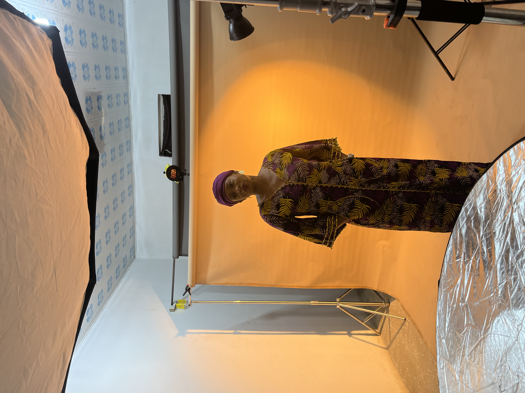 A woman wearing a purple and yellow print dress and a purple headwrap is standing in front of an orange backdrop in a photography studio. The studio features photographic lighting equipment, including a softbox to the left and a reflector to the right of the subject. The wall behind the softbox is decorated with a blue and white floral pattern. The studio location is Gura Top, Nigeria.