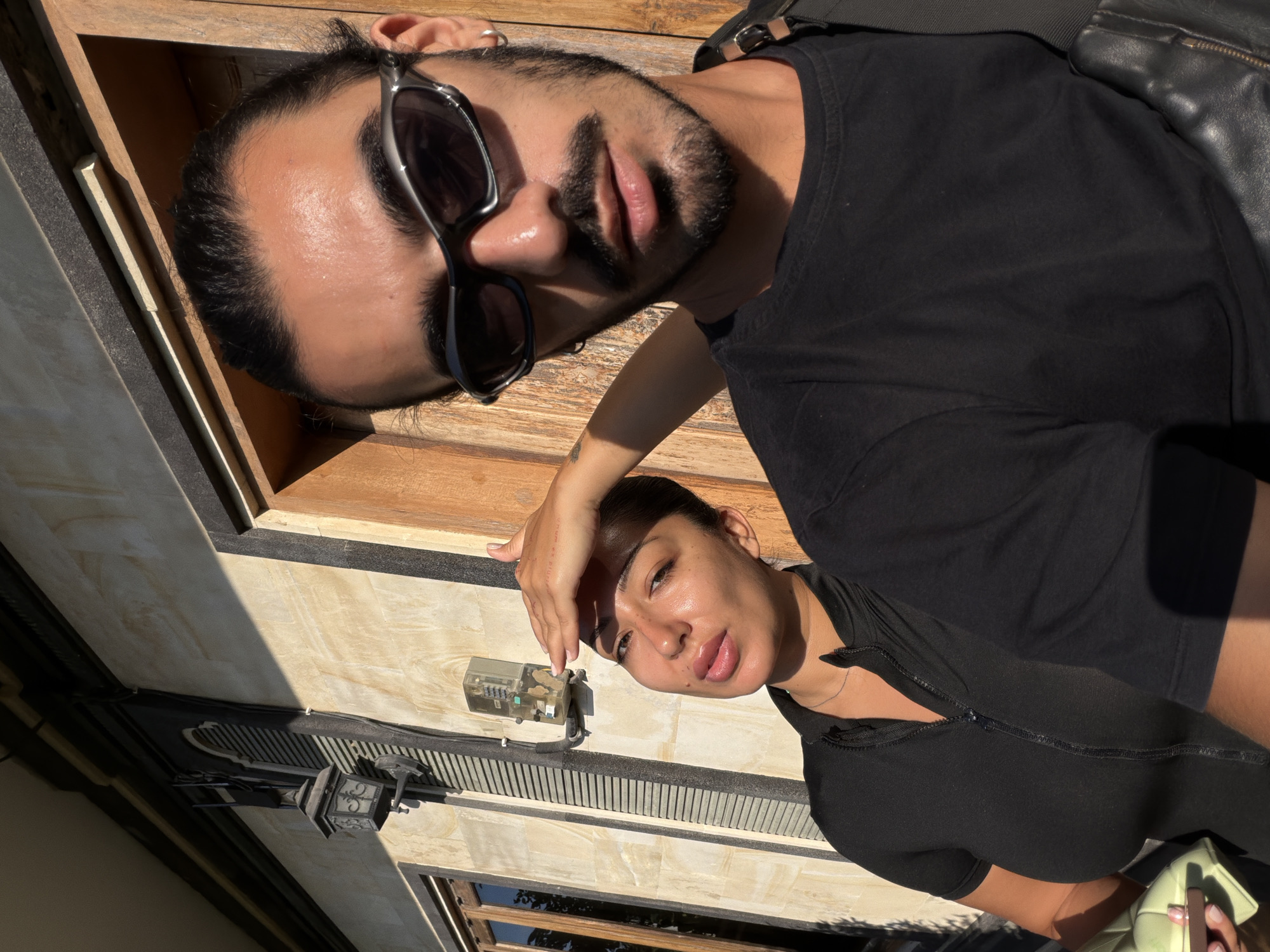 The image shows two individuals posing for a photograph in an outdoor setting. A man with a beard and sunglasses is positioned at the top of the frame, wearing a black shirt and a leather jacket. A woman with her hair pulled back is positioned below him, shielding her eyes with her hand. Both individuals are facing the camera.
The location appears to be in North Kuta, Indonesia, based on architectural details and the style of the building facade. The background includes a light-colored wall with dark trim, and a small electrical box is visible on the wall near the woman. A vintage-style outdoor light fixture is also attached to the wall. Sunlight and shadows are present, indicating a bright, sunny day.