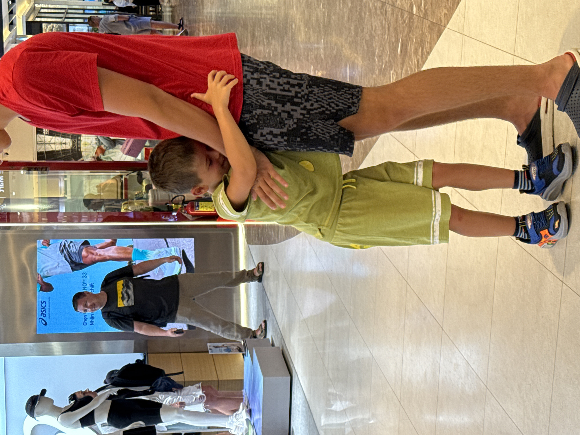 In Nha Trang Ward, Vietnam, a young boy in a green t-shirt and shorts hugs the leg of a taller person wearing a red t-shirt and patterned shorts. The boy's face is partially obscured as he presses it against the taller person's leg. Behind them, a man in a dark t-shirt and khaki pants is visible, looking to his left. To the far left, a mannequin dressed in athletic wear stands on a display platform. The environment appears to be an indoor public space, possibly a shopping mall, with a tiled floor and various displays and advertisements in the background.